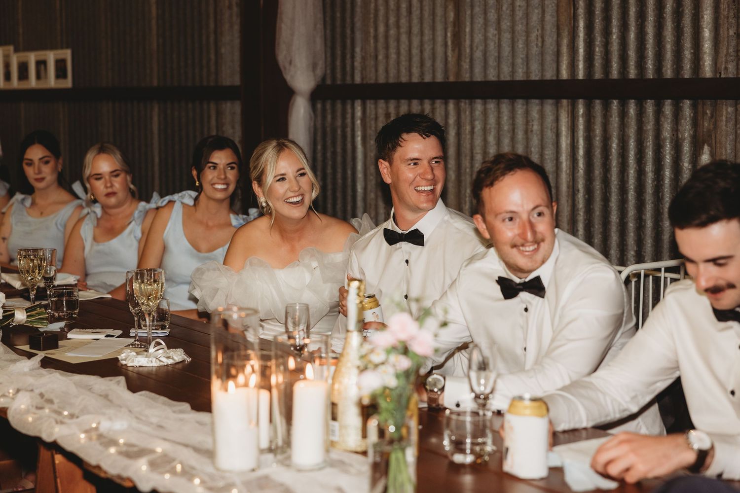 Wedding party seated at a long rustic table during reception dinner with candlelight and decorations.