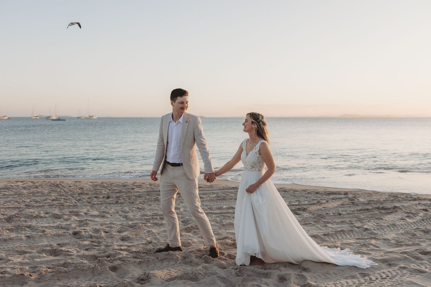 A couple walks hand in hand along a scenic beach at sunset with gentle waves and mountains in the background.