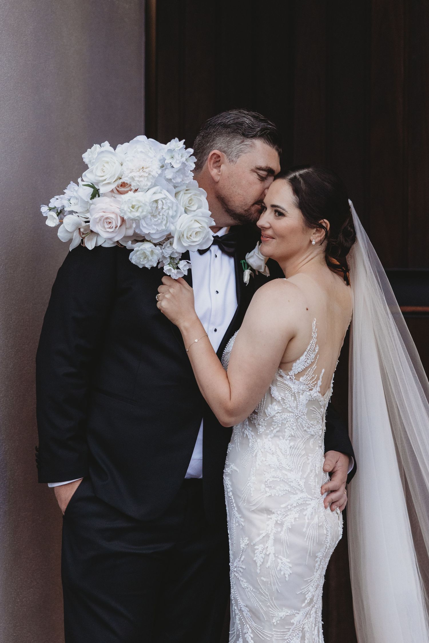 Intimate wedding couple portraits with white orchid bouquet against dark architectural backdrop.