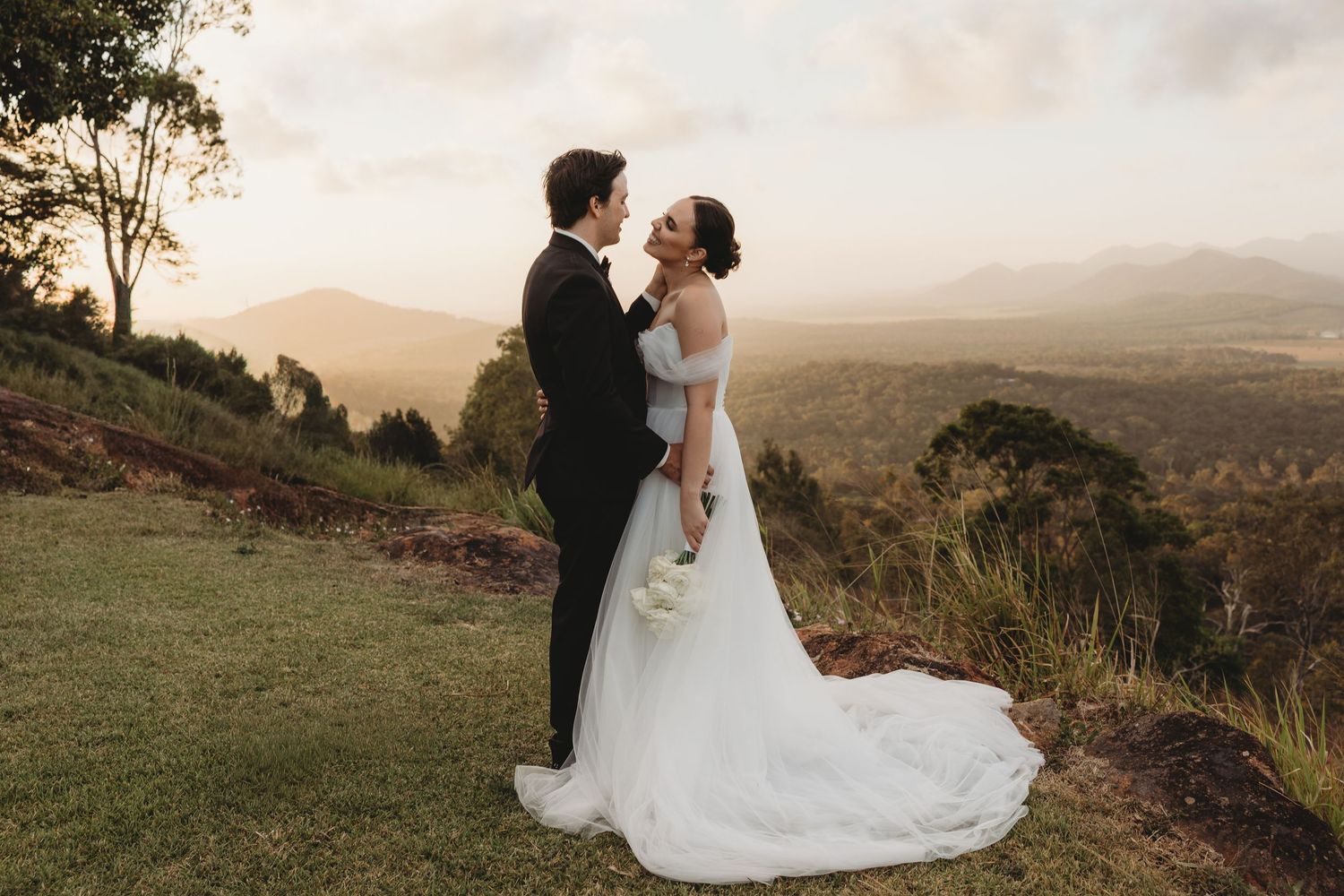 Wedding couple embraces on grassy hilltop with mountain vista in background.