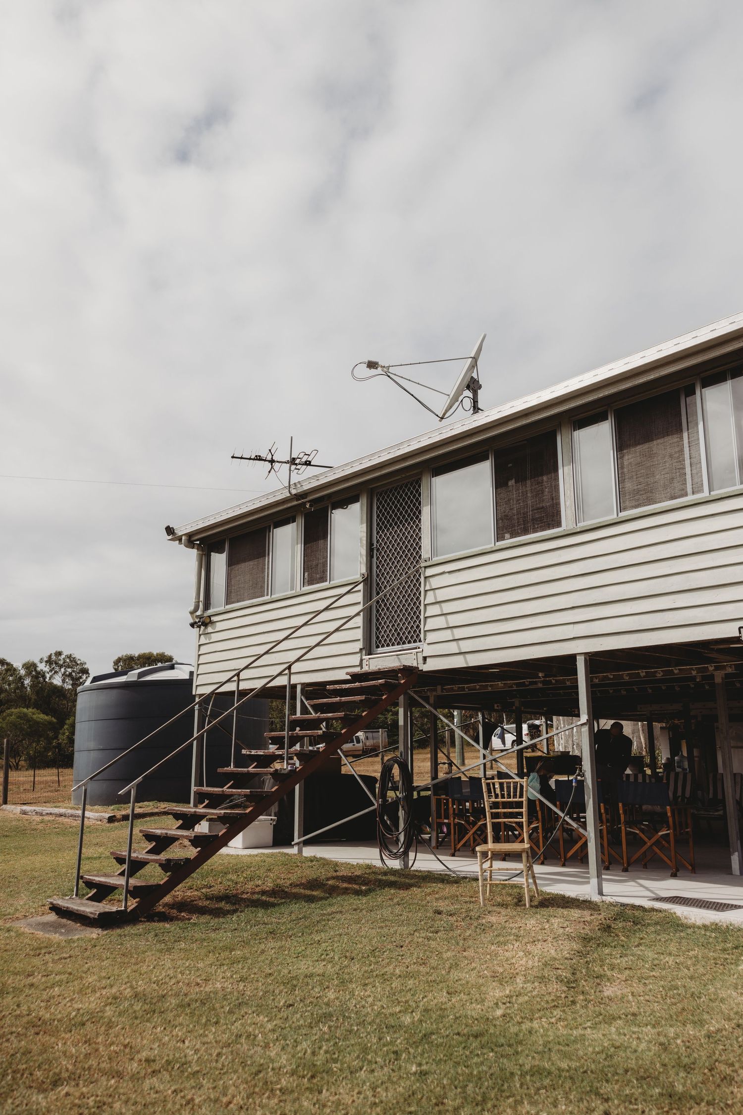 Rustic elevated farmhouse with metal staircase, water tank, and satellite antenna under cloudy sky.