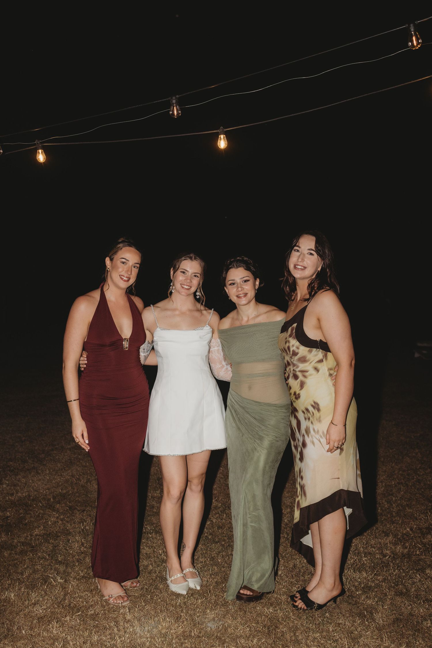 Bride poses with three friends under string lights at nighttime outdoor wedding reception.