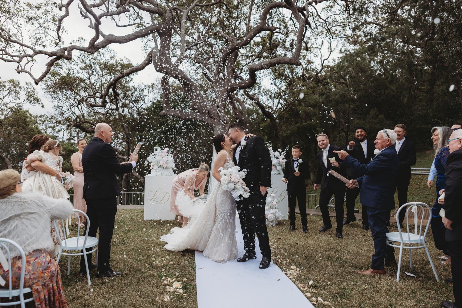 A romantic outdoor wedding ceremony takes place under blooming trees with guests seated on white chairs.