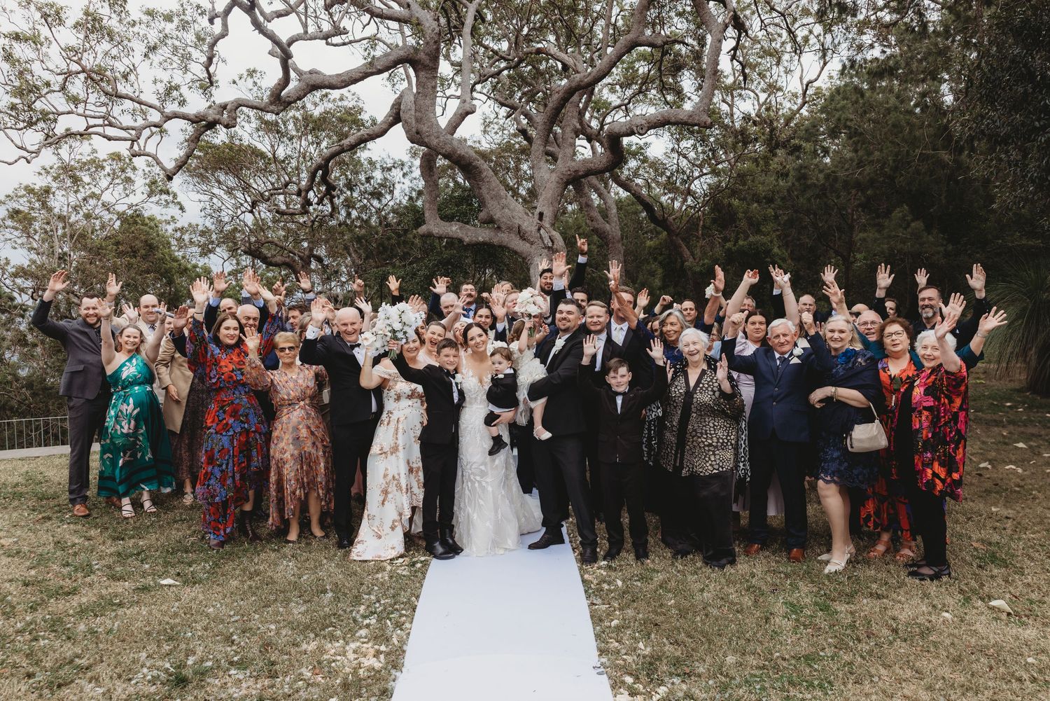 Large group wedding photo with guests gathered outdoors under trees.