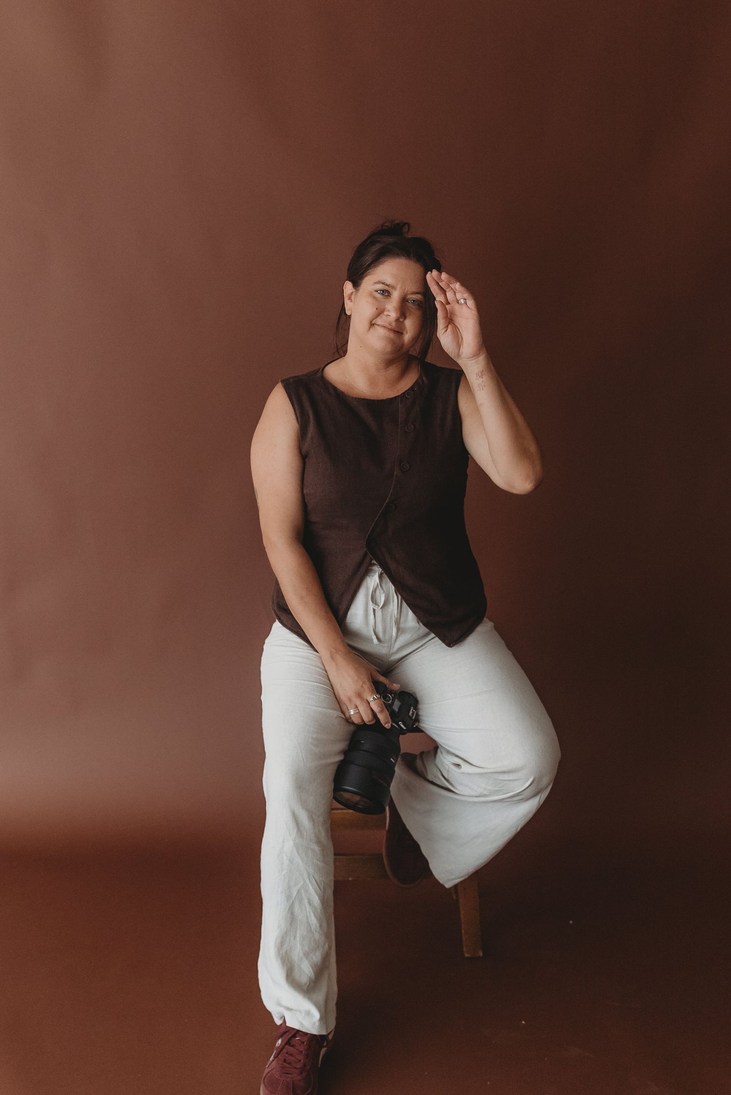 Portrait of photographer in casual attire holding camera while sitting on stool.