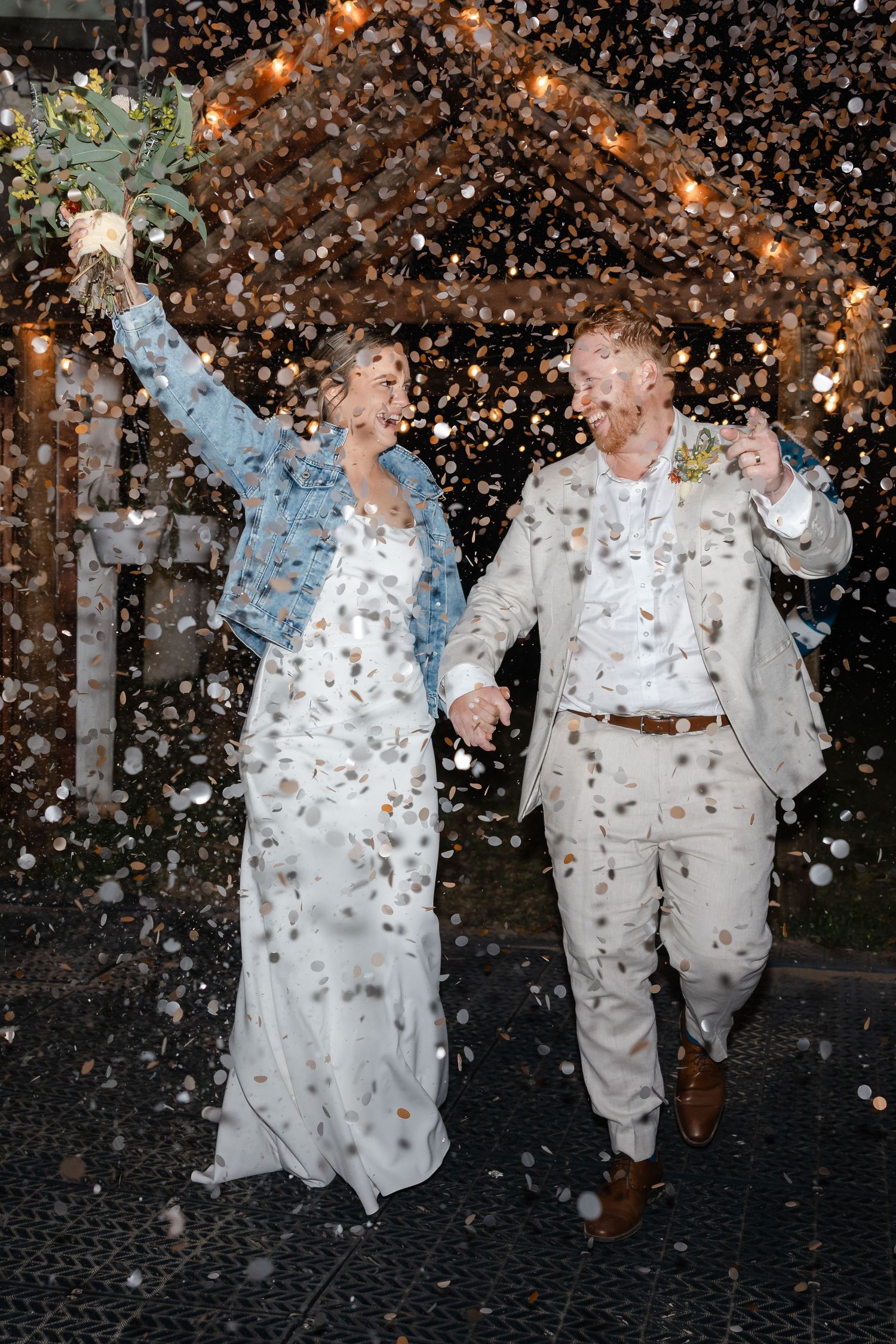 Joyful newlyweds celebrate with confetti shower, bride in white gown and denim jacket holding bouquet aloft.