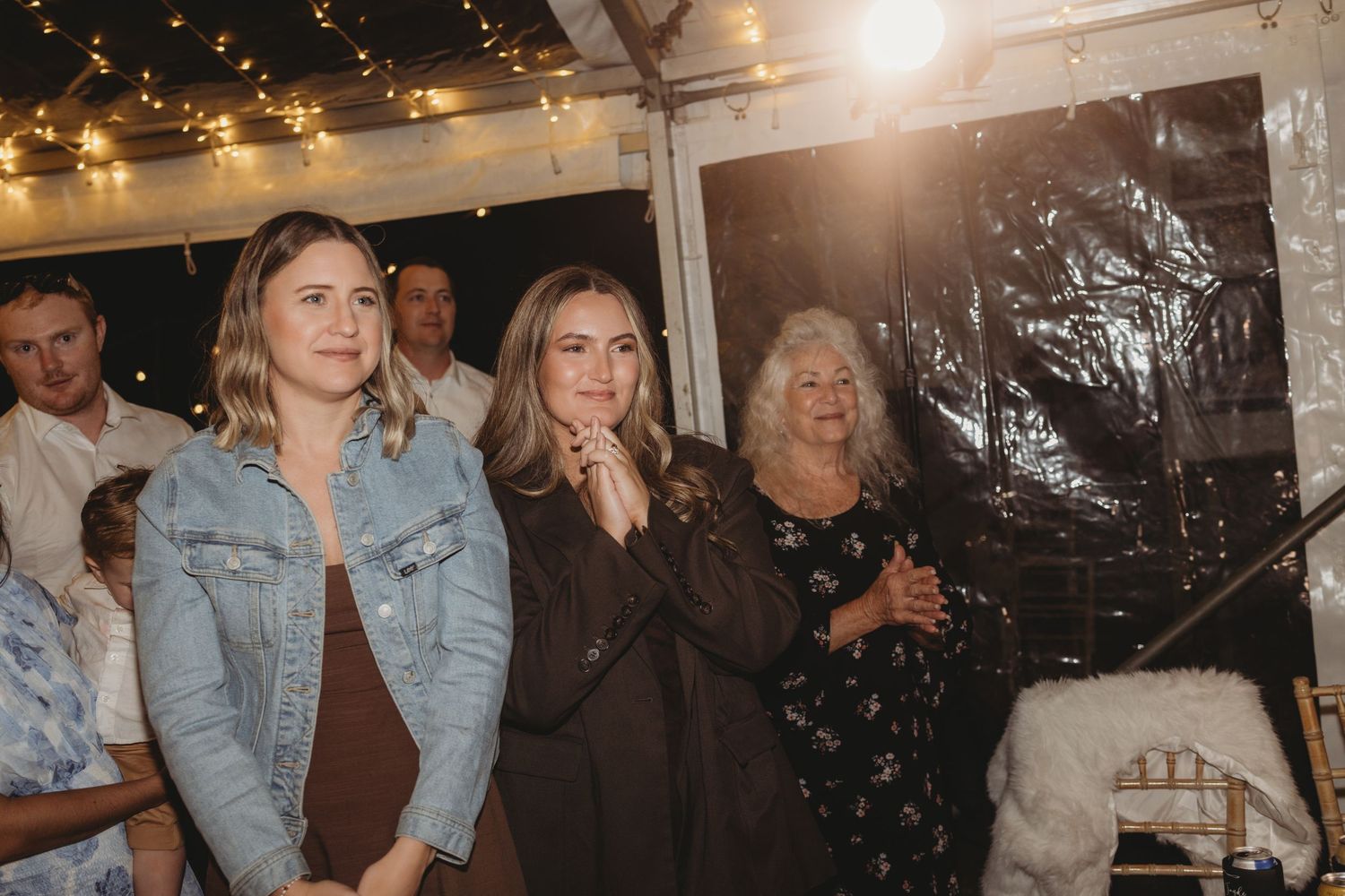 Three women stand together watching an emotional moment at an indoor wedding reception under festive lights.