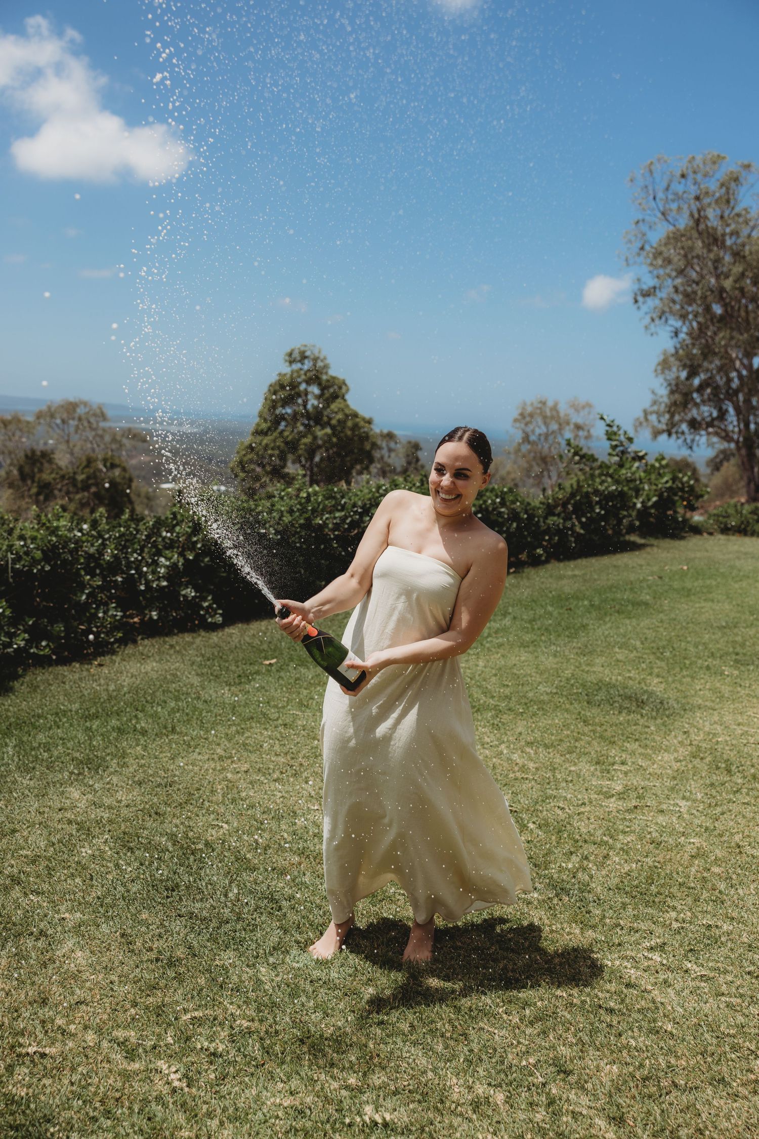 Someone in a white dress sprays champagne on a sunny grassy hillside.