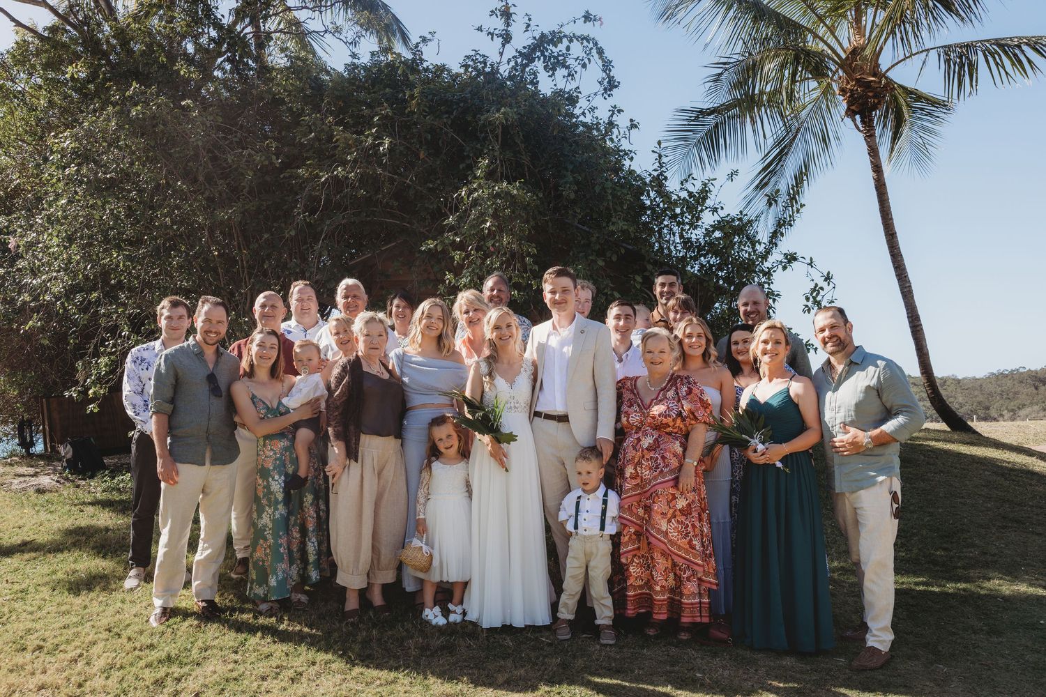 Wedding party group photo with guests gathered outdoors under palm trees.