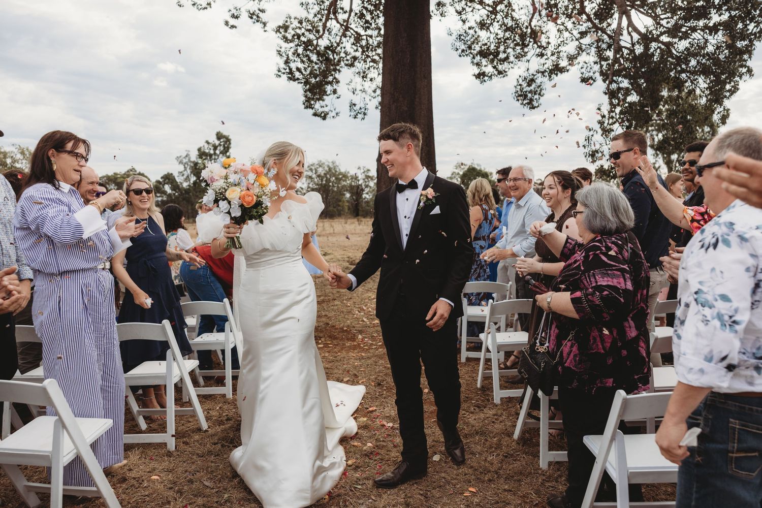 Wedding ceremony recessional with guests seated on white chairs beneath a towering tree.