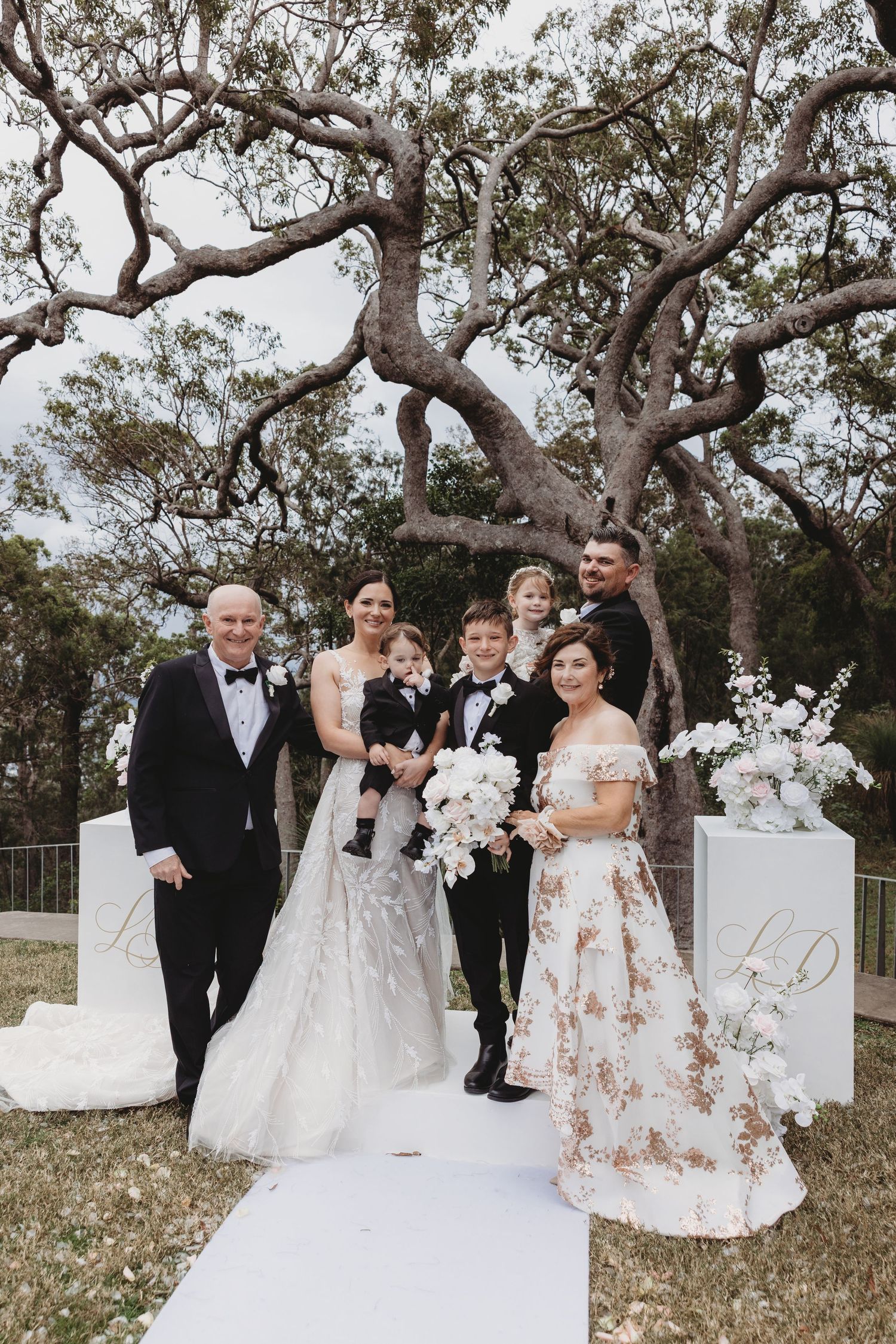 Wedding party members pose together under a large tree with white floral decorations.