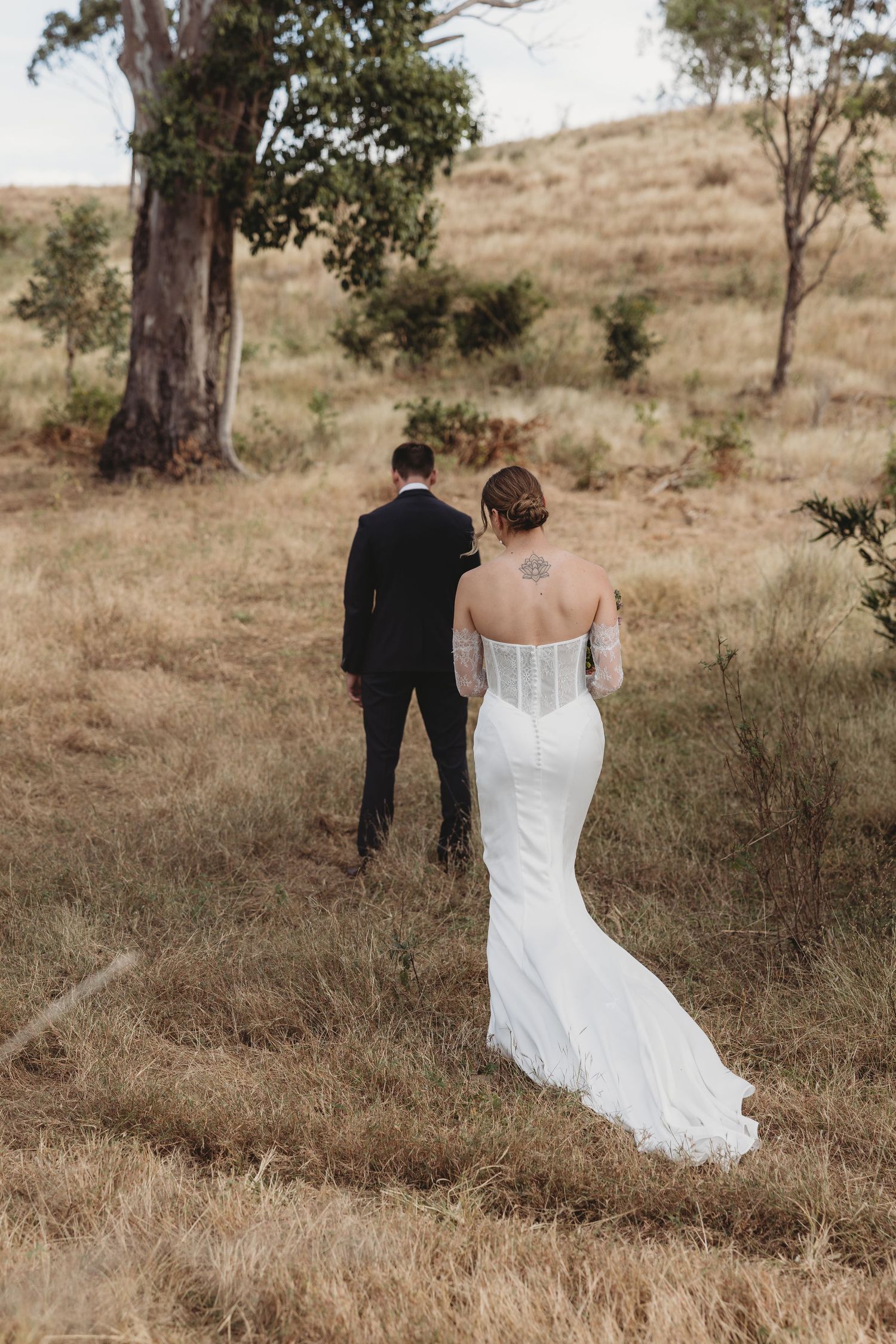 Bride and groom walking through golden grass fields with eucalyptus trees, bride wearing elegant off-shoulder mermaid gown.
