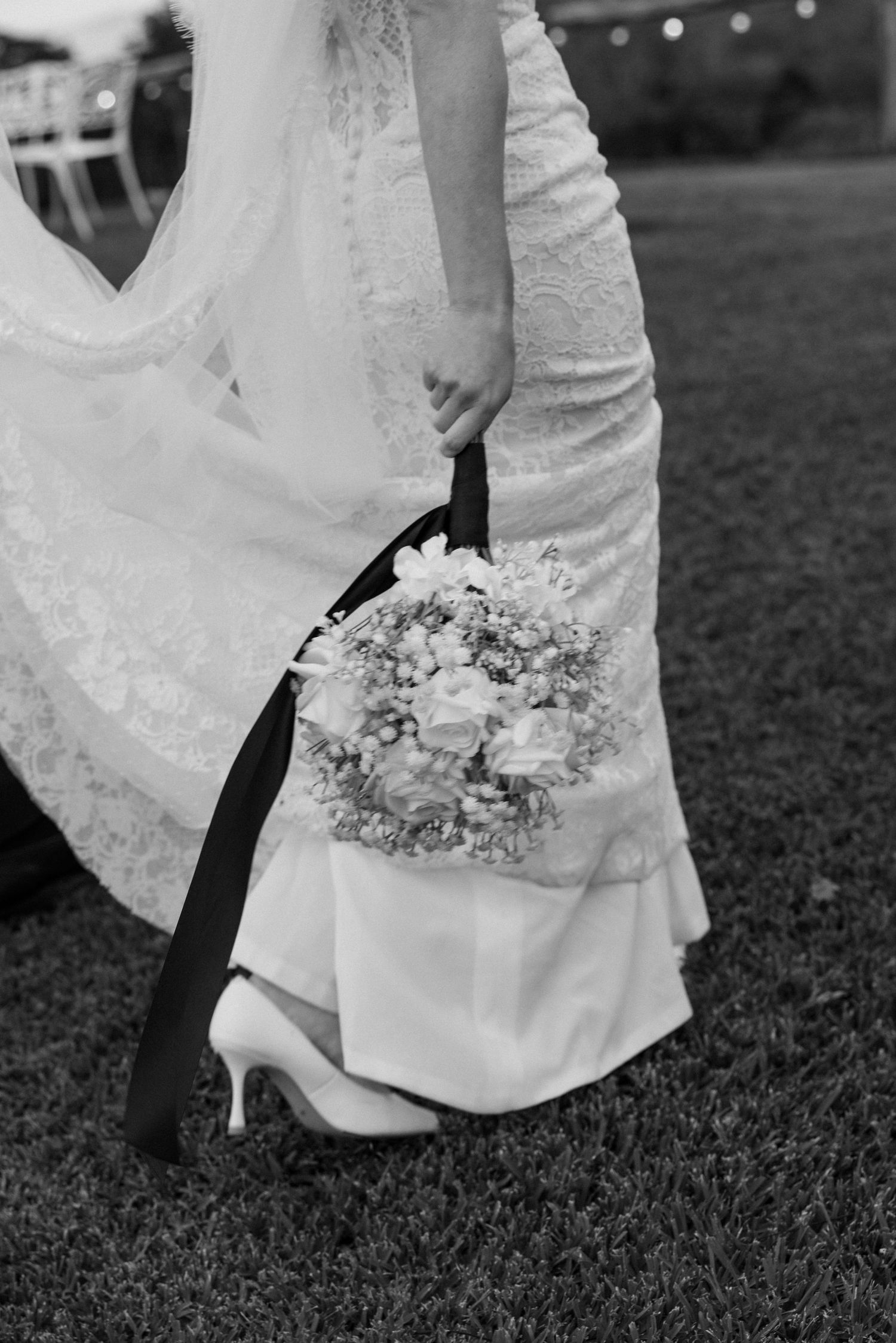 Black and white photo of a bride in lace gown holding a floral bouquet, wearing heels on a grass lawn.