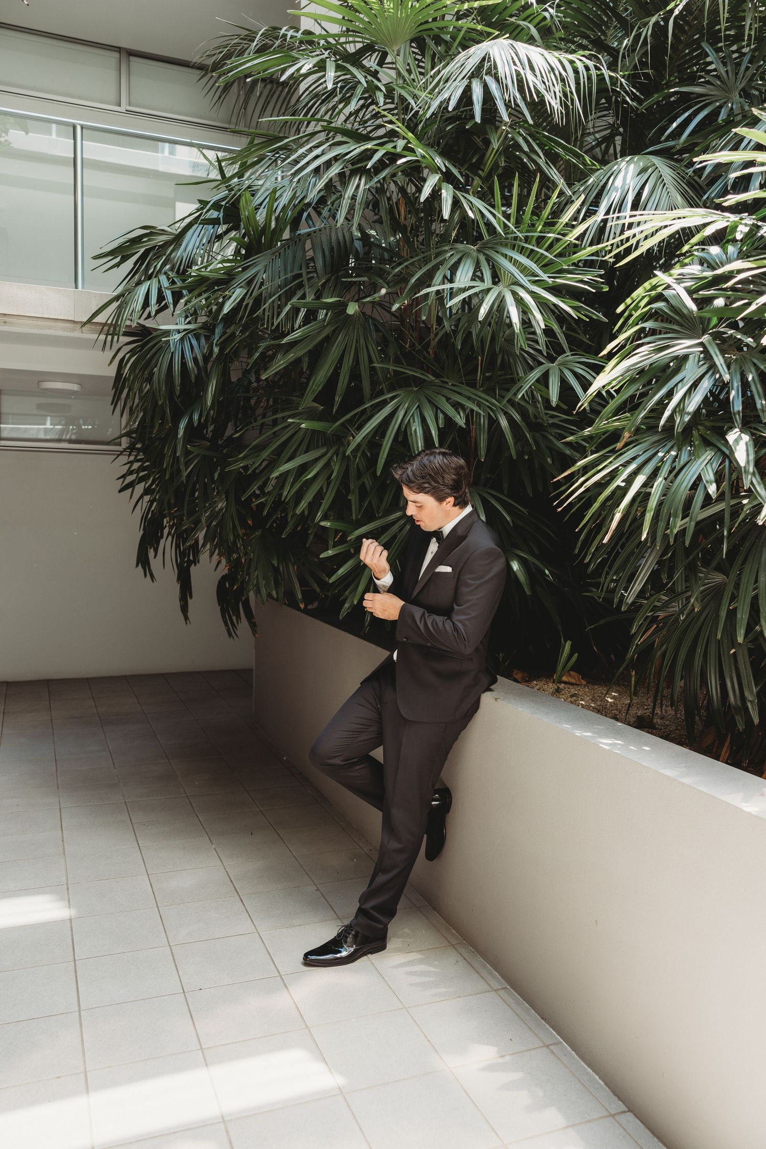 A person in a tuxedo sits thoughtfully by tropical plants in an indoor setting.