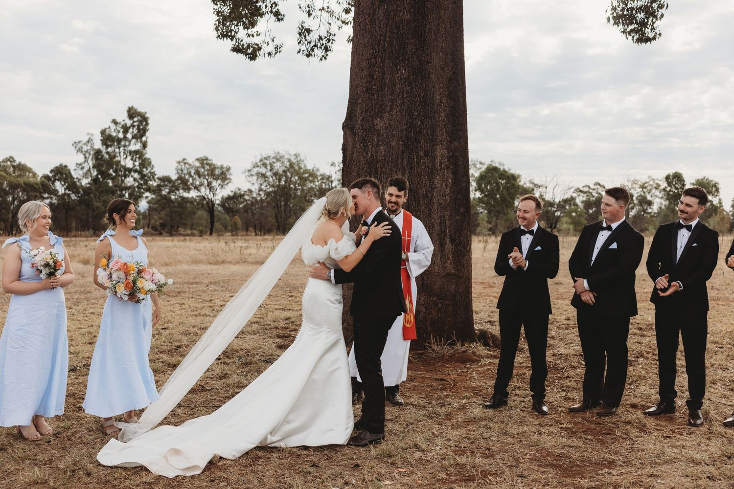 Wedding party poses beneath tall trees in rustic outdoor setting with bridesmaids in blue and groomsmen in black.