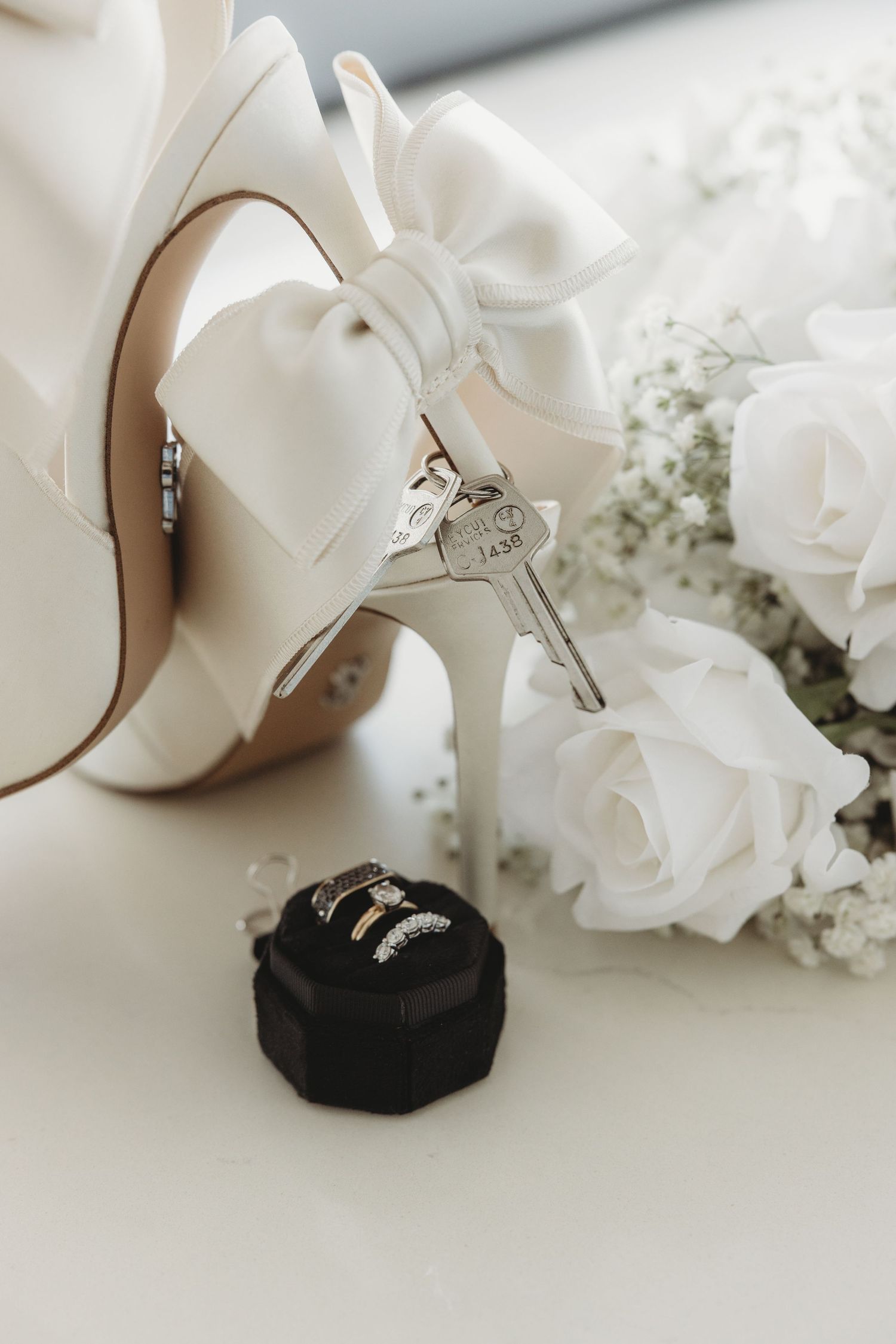 Elegant white bridal shoe with bow detail next to roses and a black ring box on white surface.