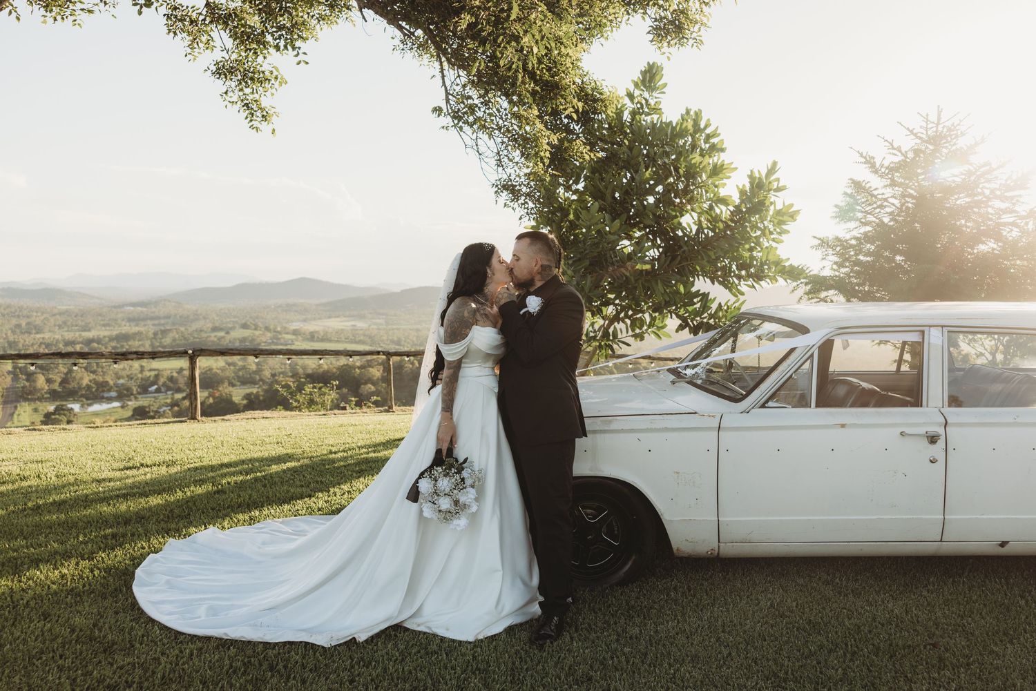 Scenic wedding photo shoot with vintage white car and sunset lighting in countryside setting.