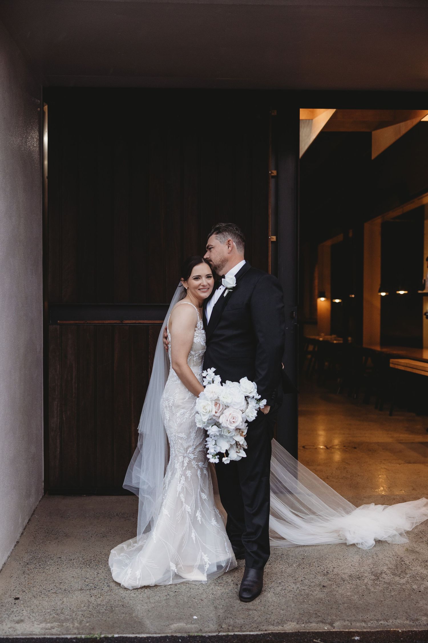 Wedding couple shares intimate moment under covered walkway in evening light.