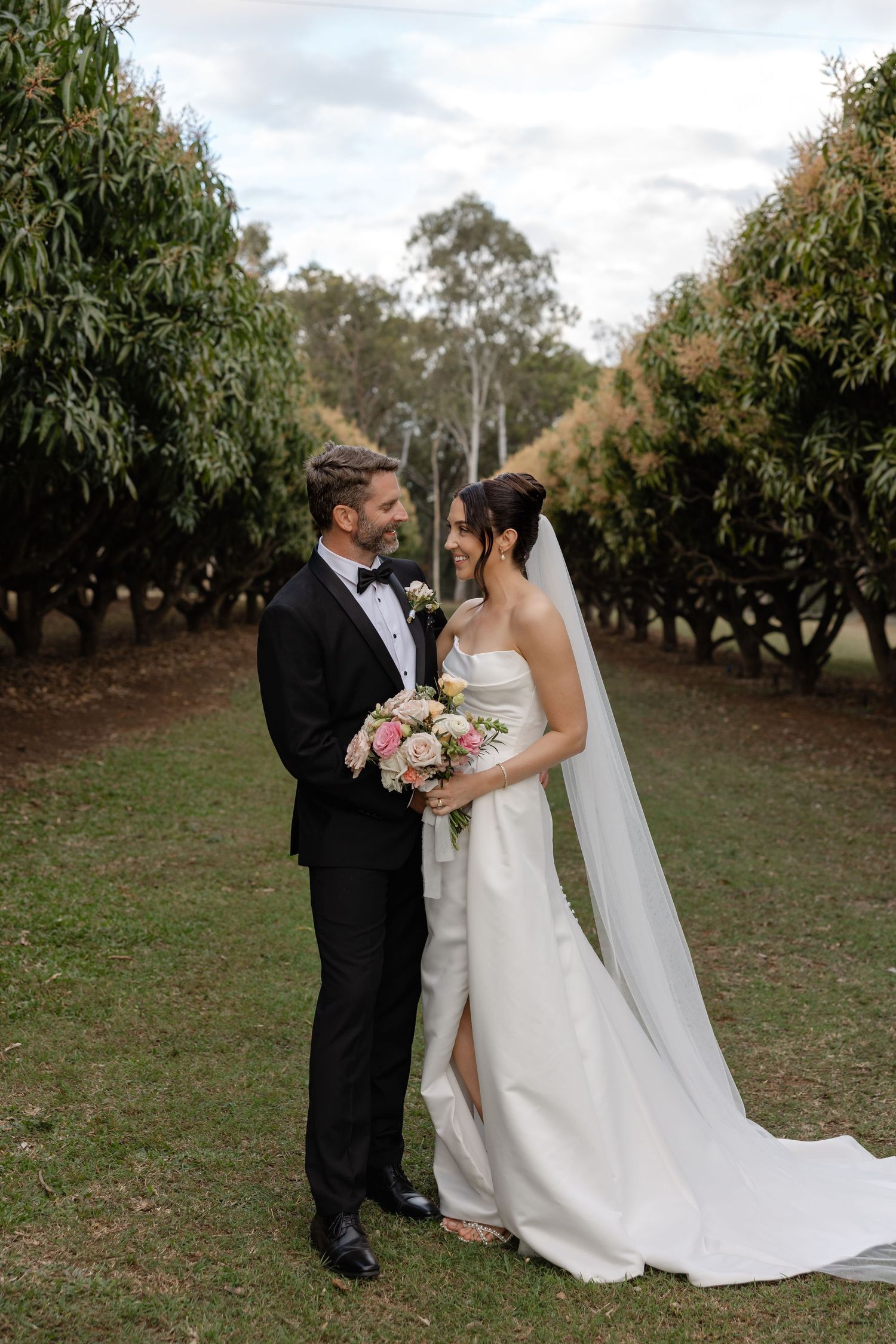 Bride and groom stand together in an orchard, bride holding a pink bouquet, wearing a white gown with veil.