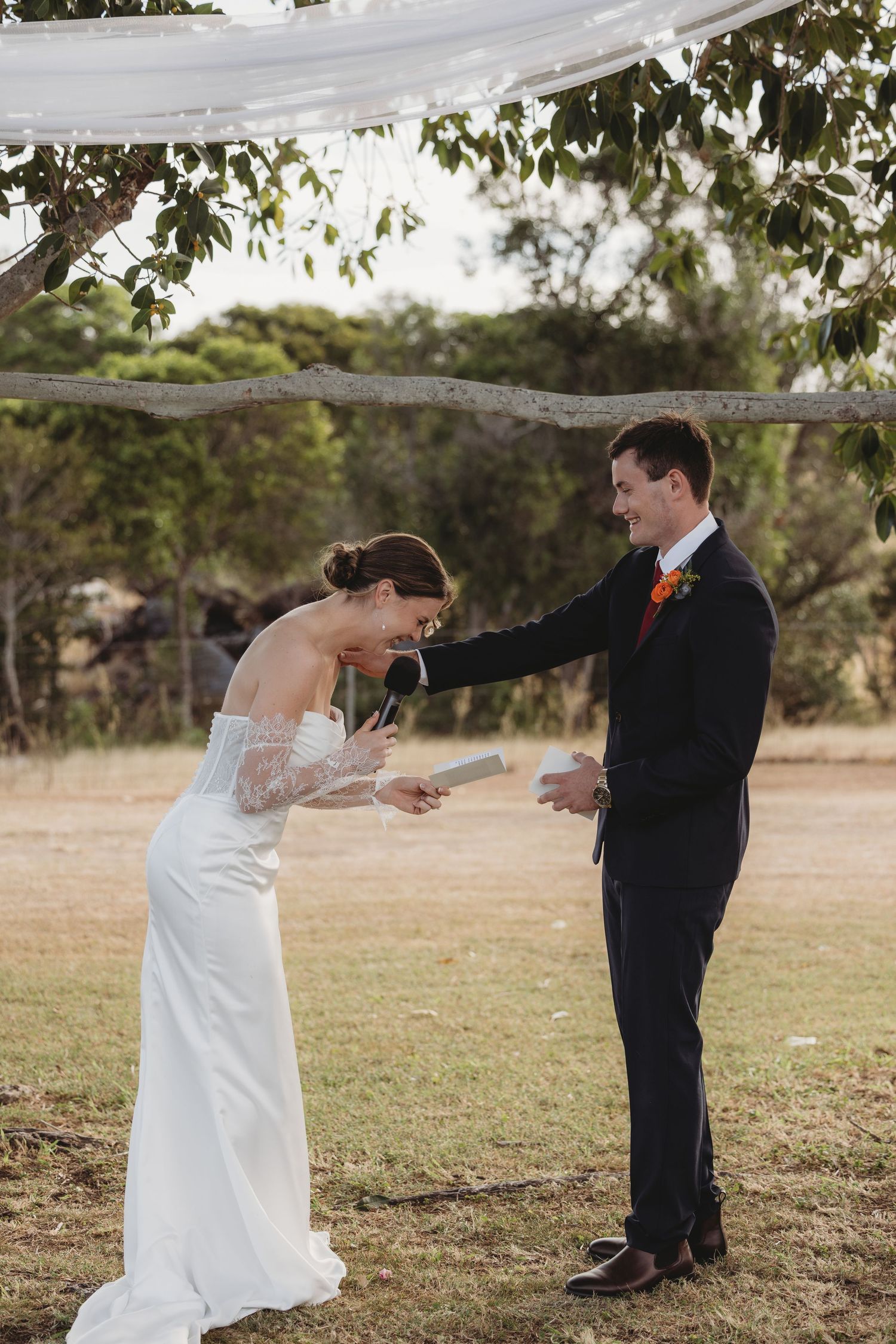 A bride in a white gown laughs while exchanging vows with her groom at an outdoor wedding ceremony.
