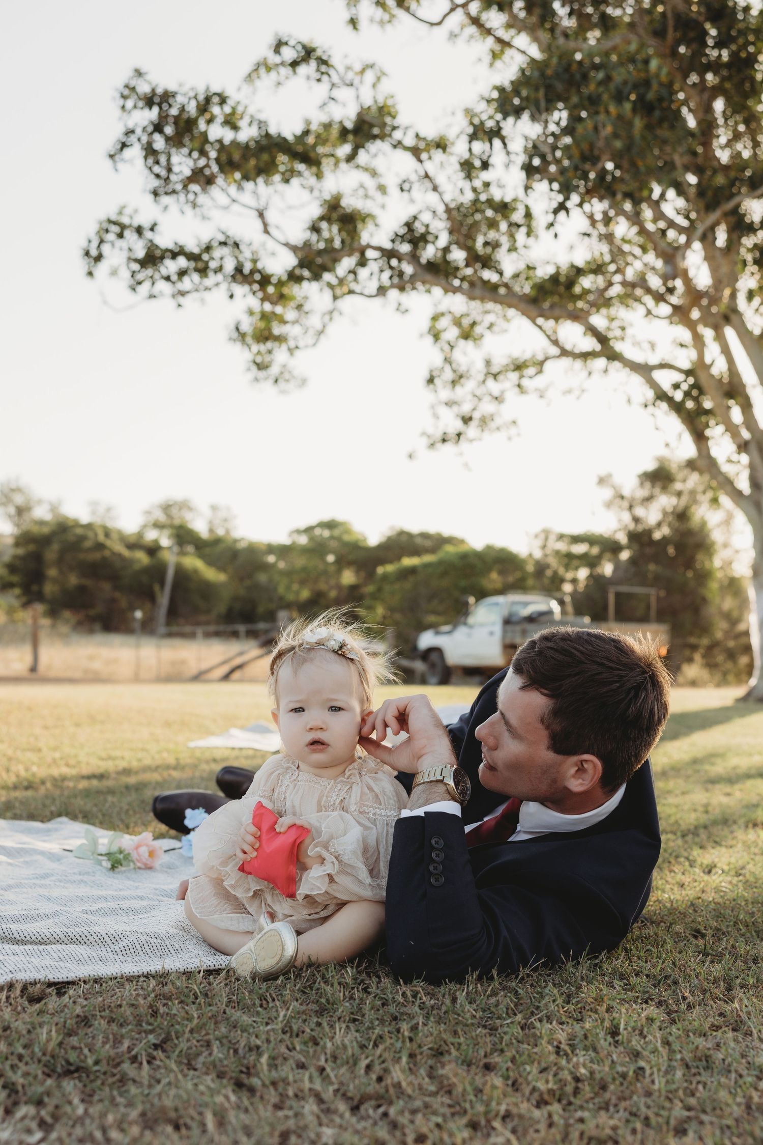 A man in a suit lies on a blanket on the grass tenderly interacting with a baby in a floral outfit.