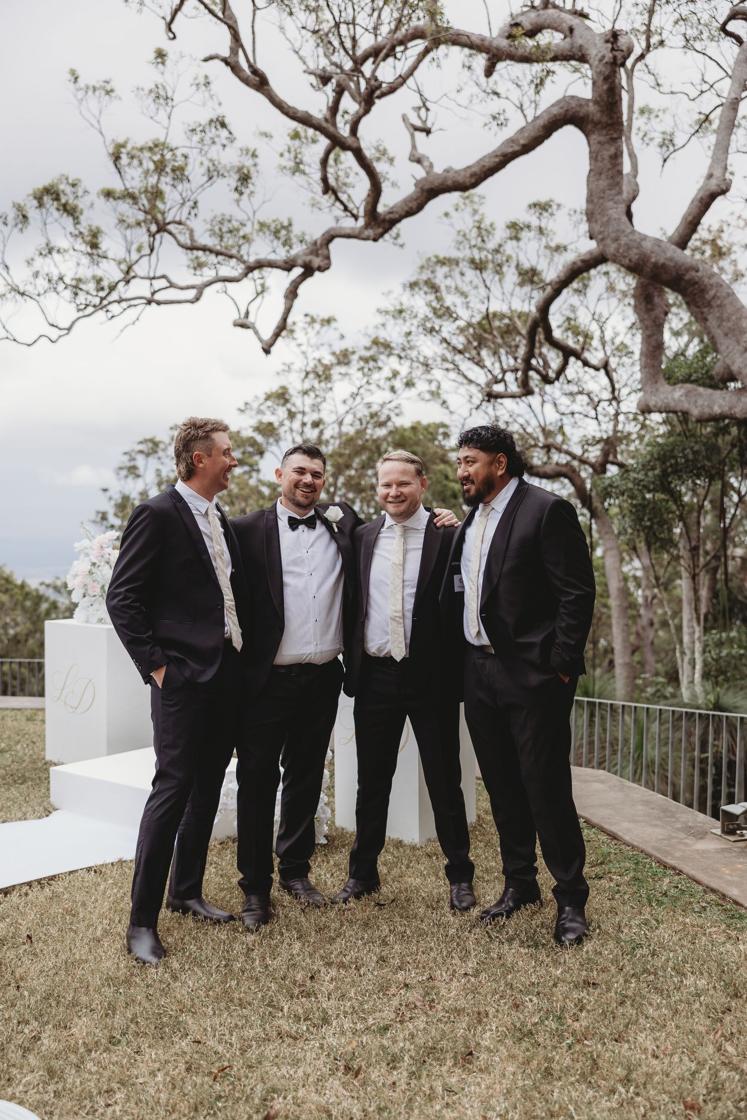 A group of groomsmen in formal attire share a laugh together under a large tree at an outdoor venue.
