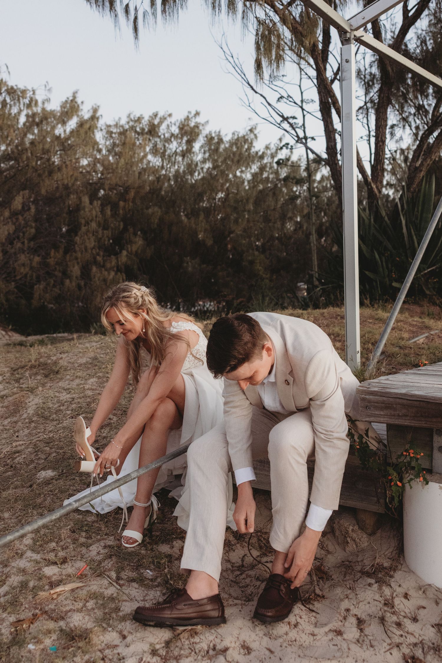 Couple in wedding attire puts on their shoes while sitting on sandy beach.