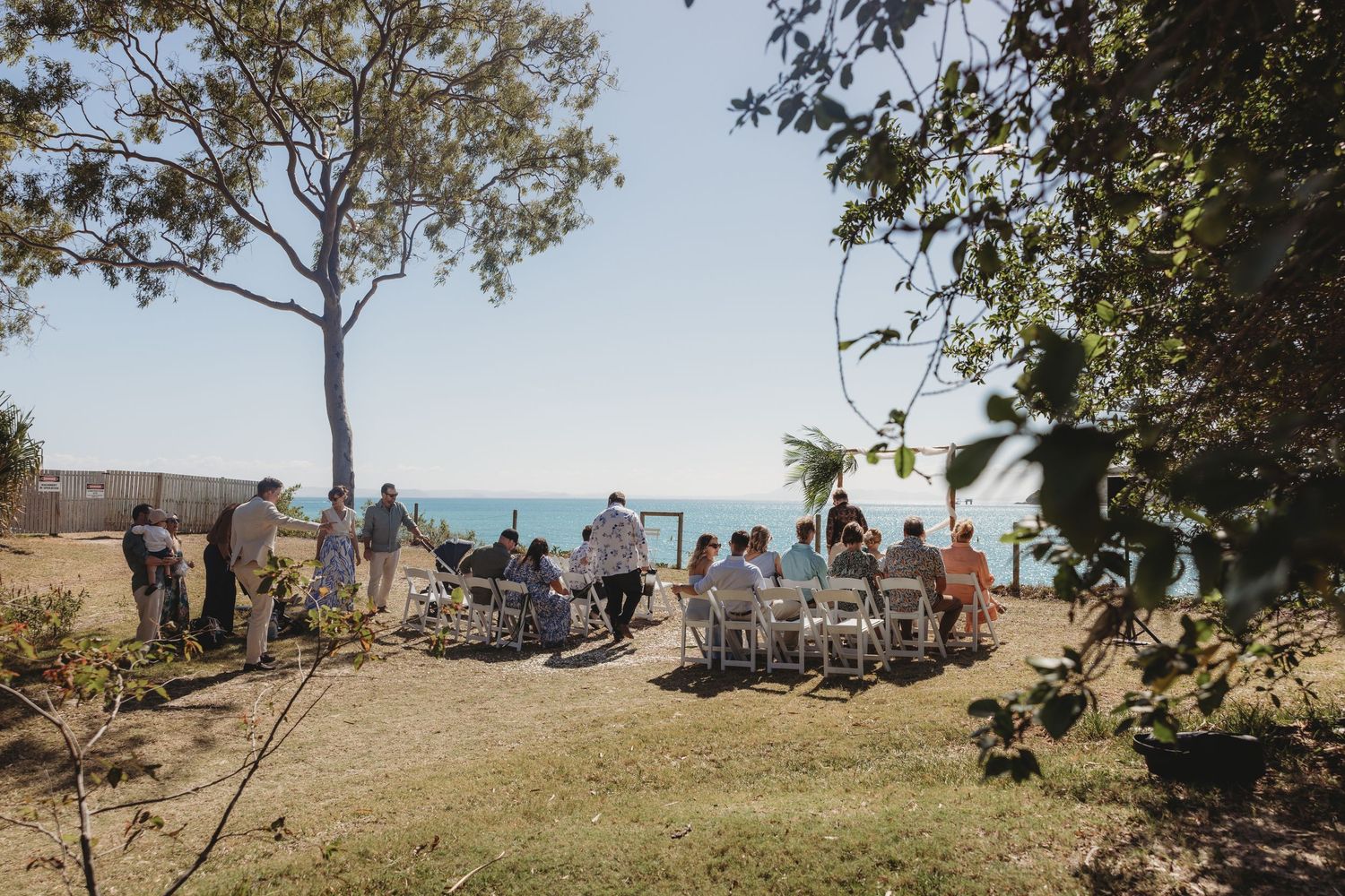 Wedding guests gathered under trees at an outdoor ceremony overlooking the ocean.