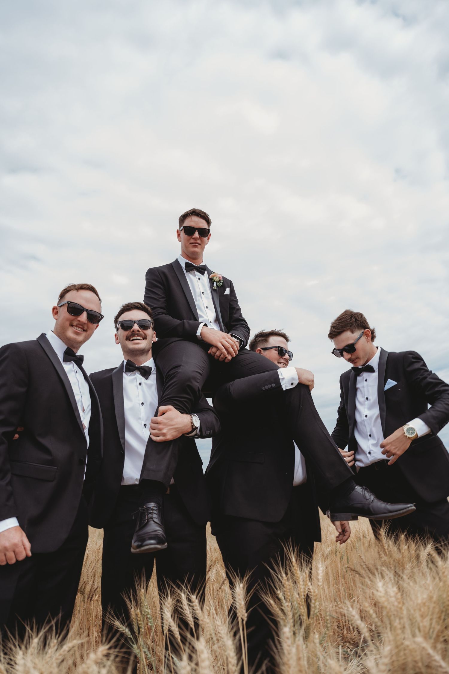 Groomsmen in sunglasses pose dramatically in golden wheat field.
