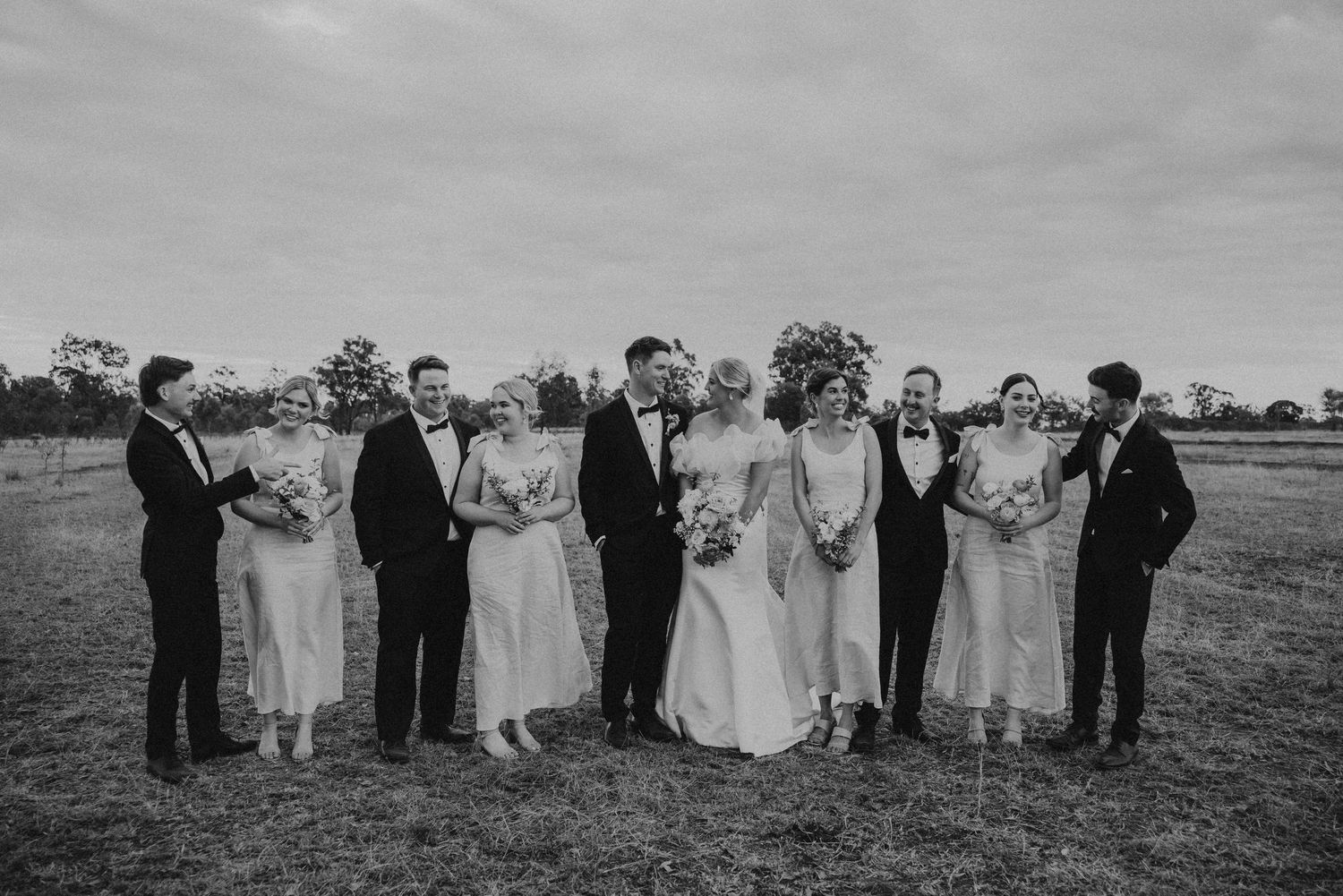 A large wedding party lines up in formation across a rural field for a group photo.