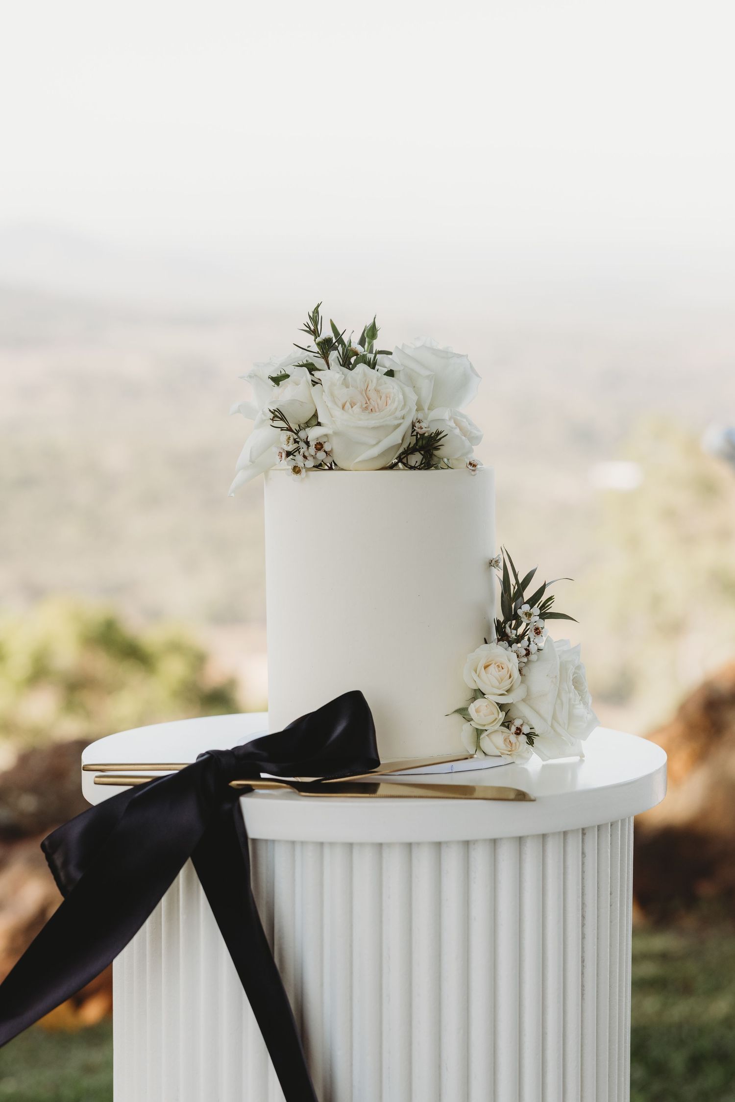 Elegant white wedding cake decorated with fresh flowers and black ribbon on a fluted pedestal.