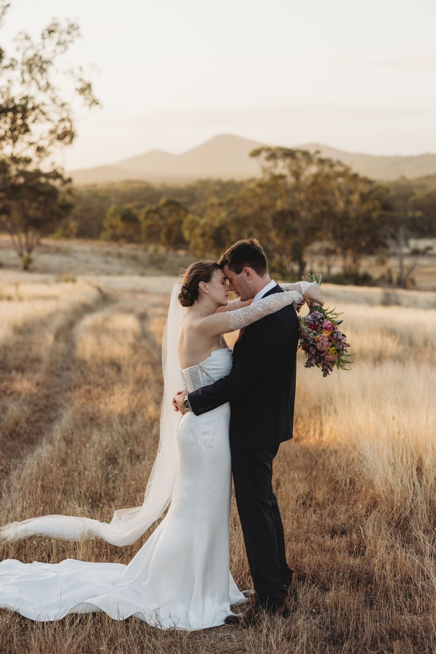Bride and groom embracing in golden field at sunset with mountain backdrop.