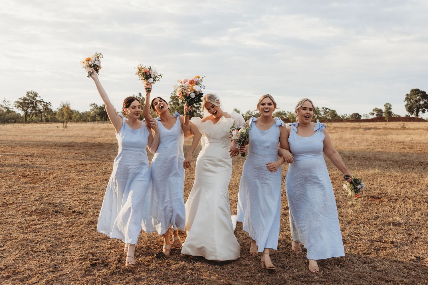Bridesmaids in light blue dresses celebrate joyfully with the bride in an outdoor field setting.