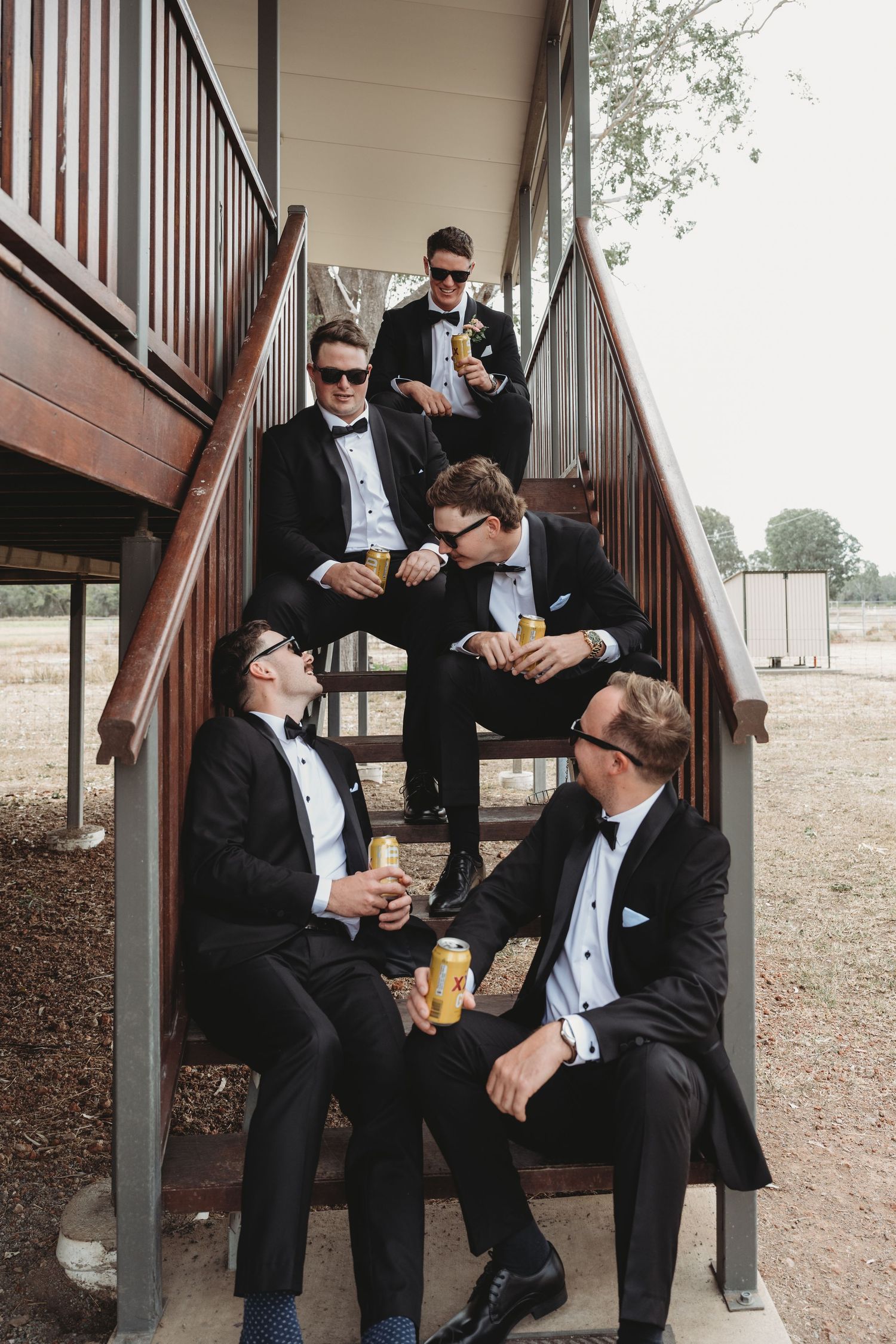 Groomsmen relaxing and sharing drinks on outdoor stairs.