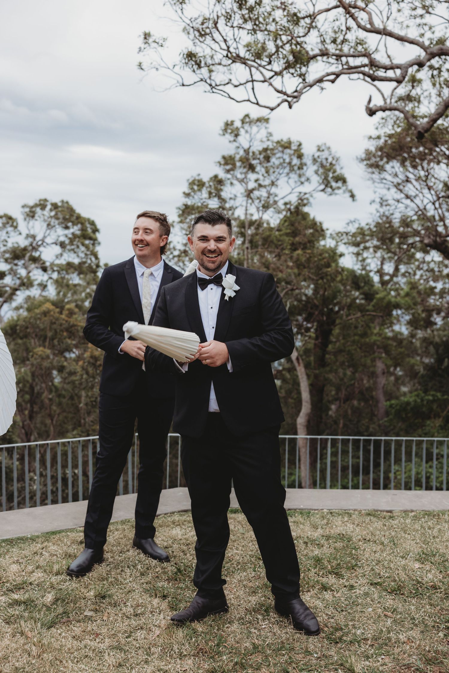 Groomsmen in black suits share laughs while sitting on grass during outdoor wedding photos.