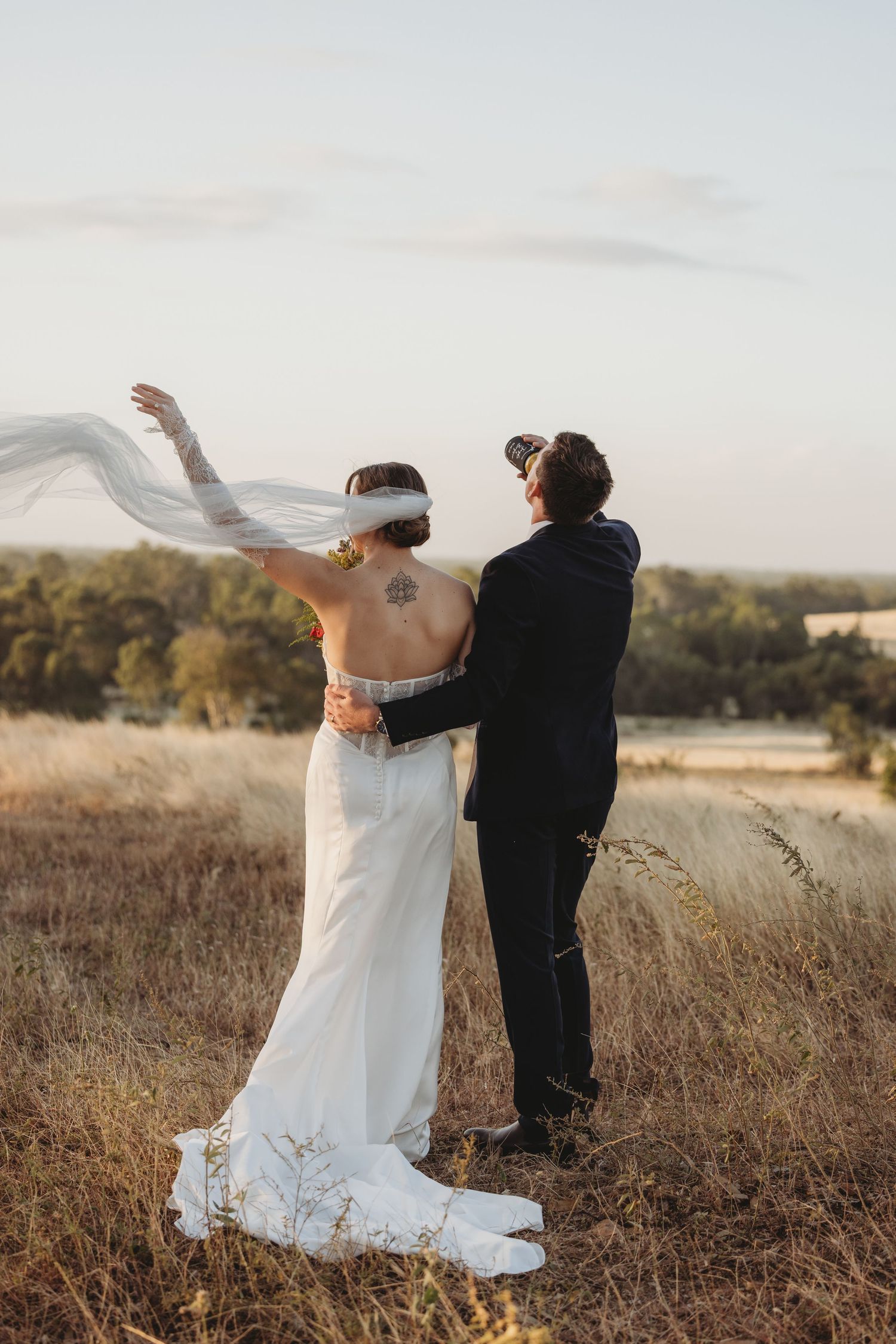 Bride and groom stand in golden field at sunset as wedding veil blows dramatically in wind.
