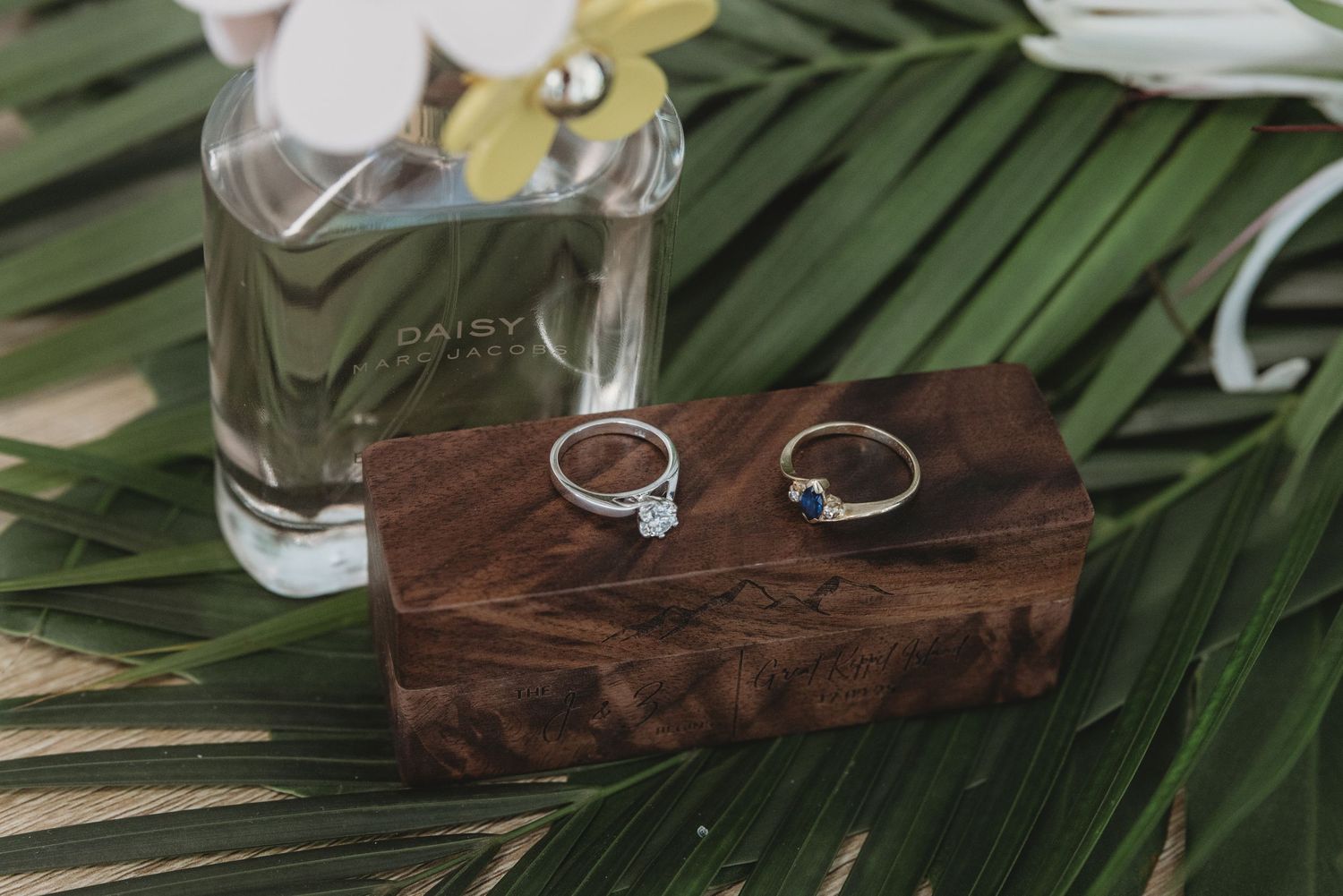 Wedding rings and jewelry displayed on a dark wooden surface with tropical palm leaves.