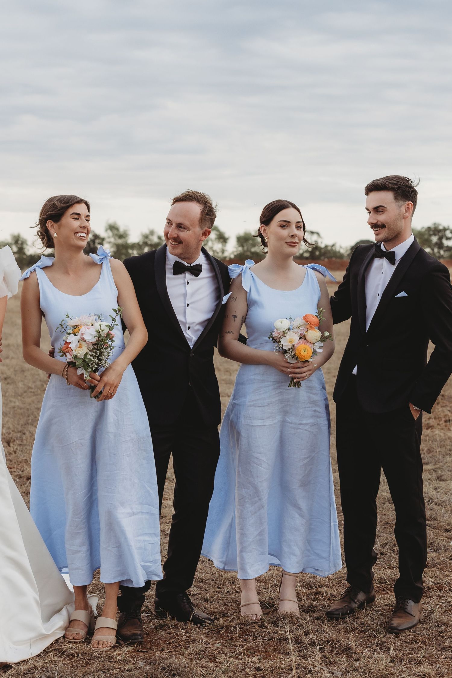 Bridesmaids in pale blue dresses and groomsmen in tuxedos laugh together on a rural landscape.