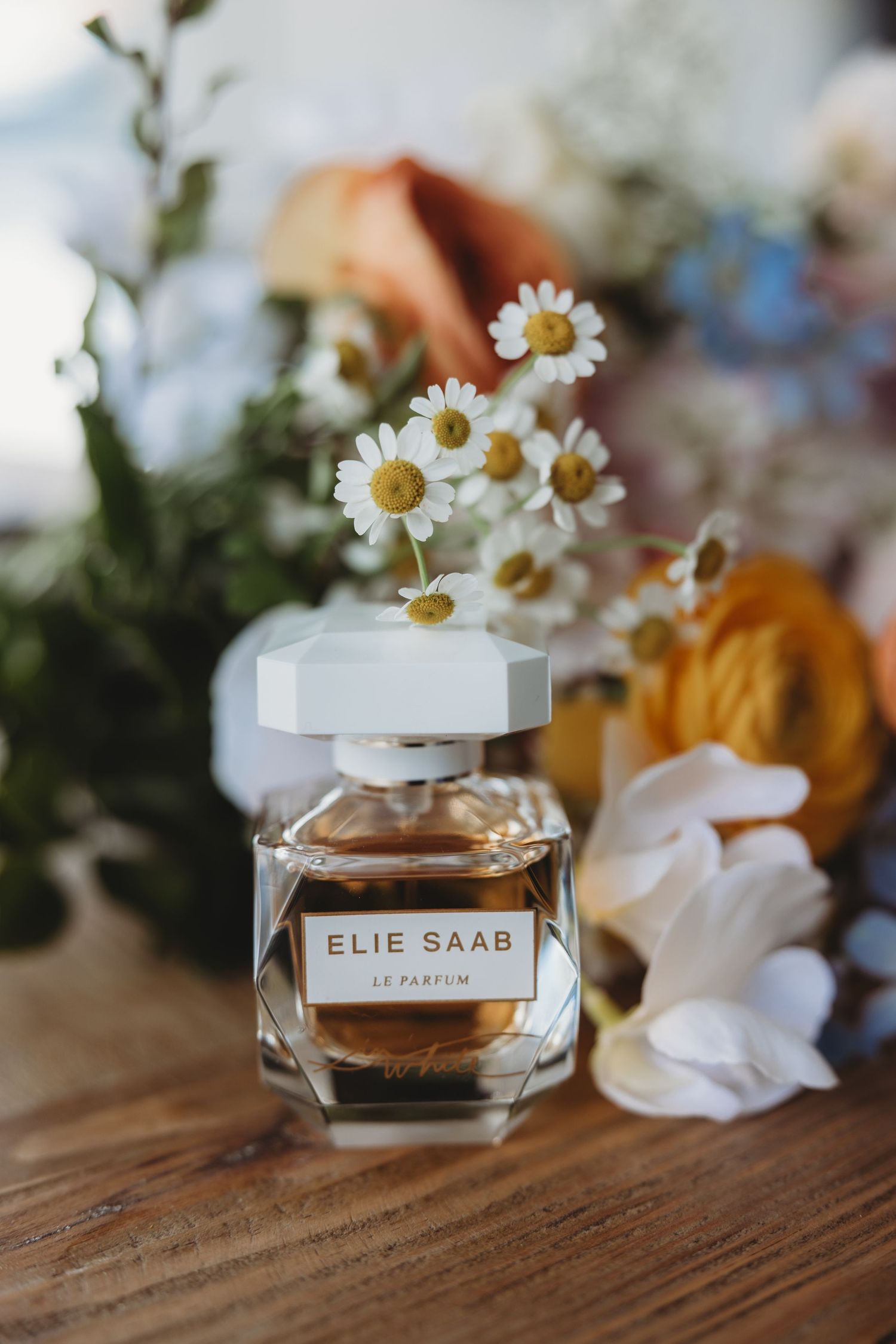 Elie Saab perfume bottle displayed with delicate white daisies and flowers on rustic wooden surface.
