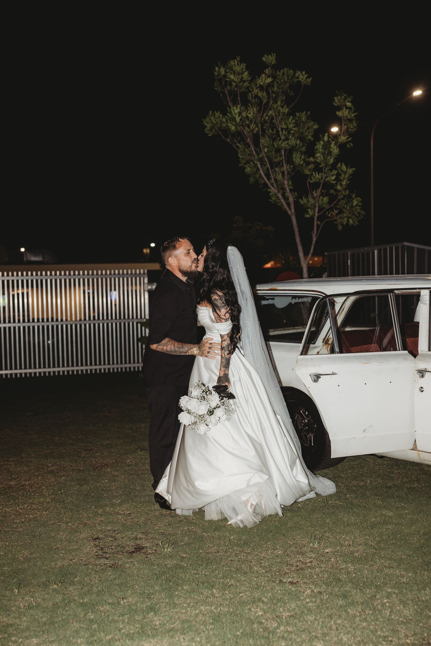 Newlyweds sharing intimate moment next to classic white car at night.