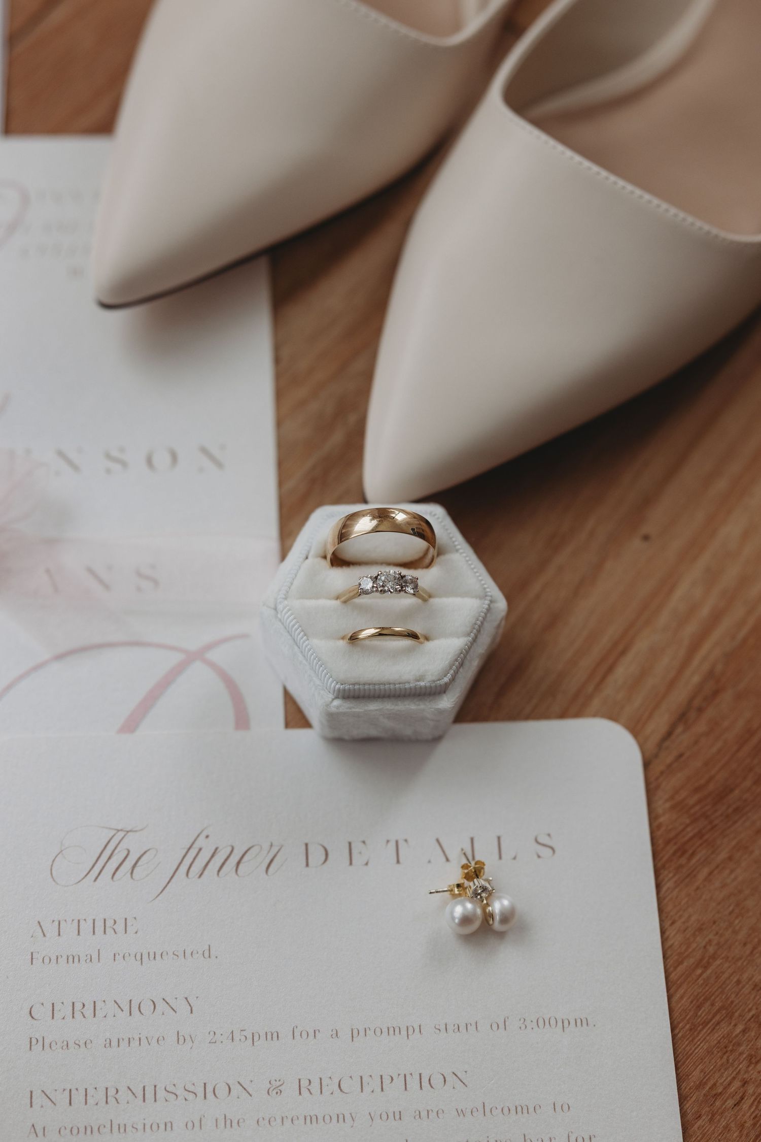 Wedding details including white heels, marble ring box, and elegant stationery on wooden surface.