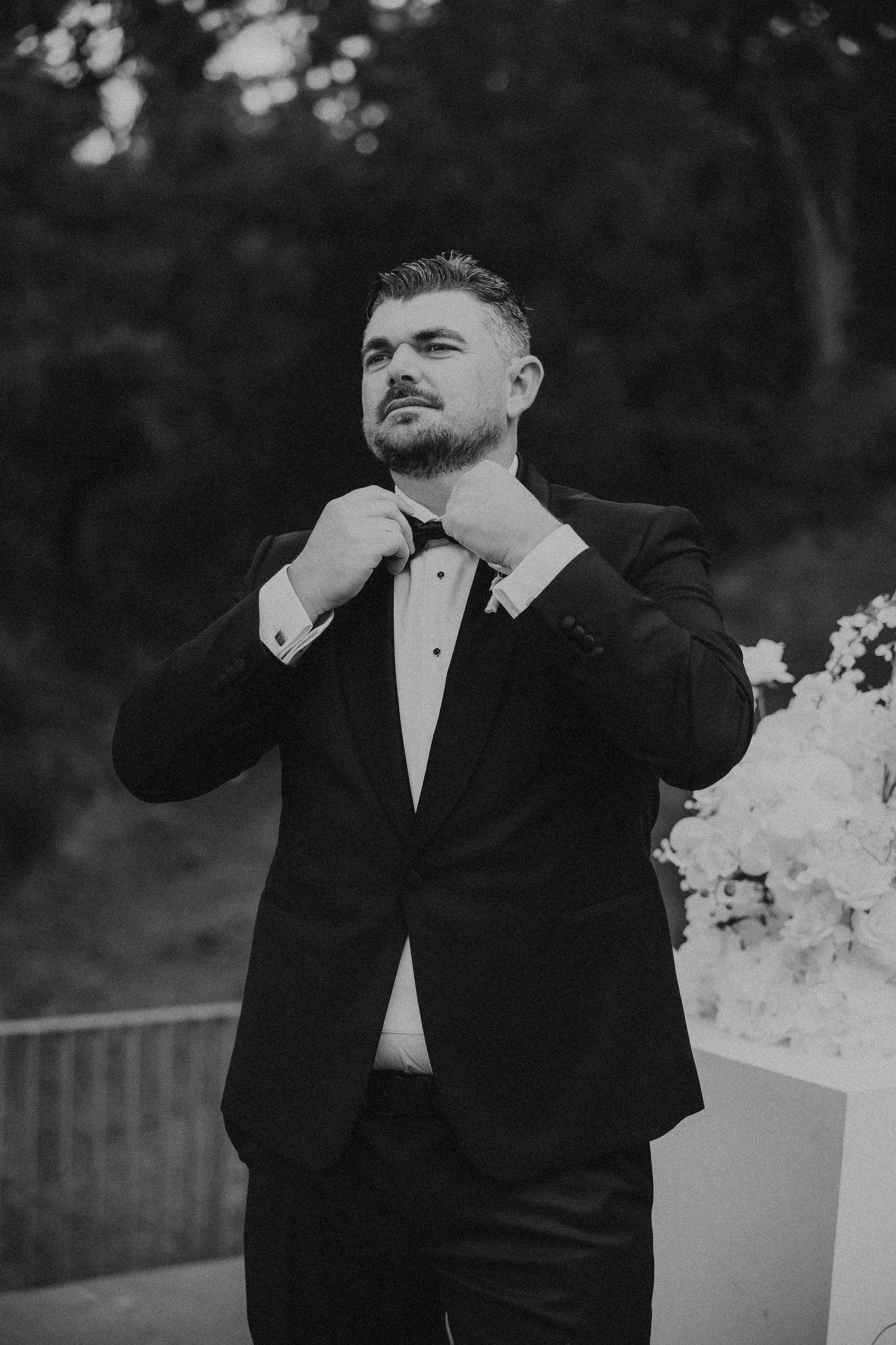 A groom in a classic black tuxedo adjusts his bowtie while standing outdoors in a black and white photo.