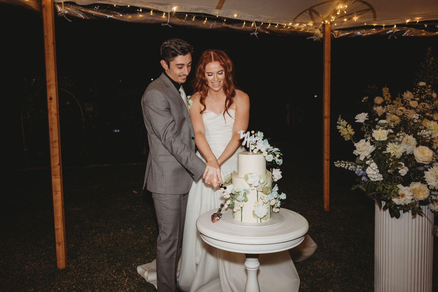 Wedding reception setup with string lights and floral arrangements creating a romantic evening atmosphere. TMF cakes St aubins wedding