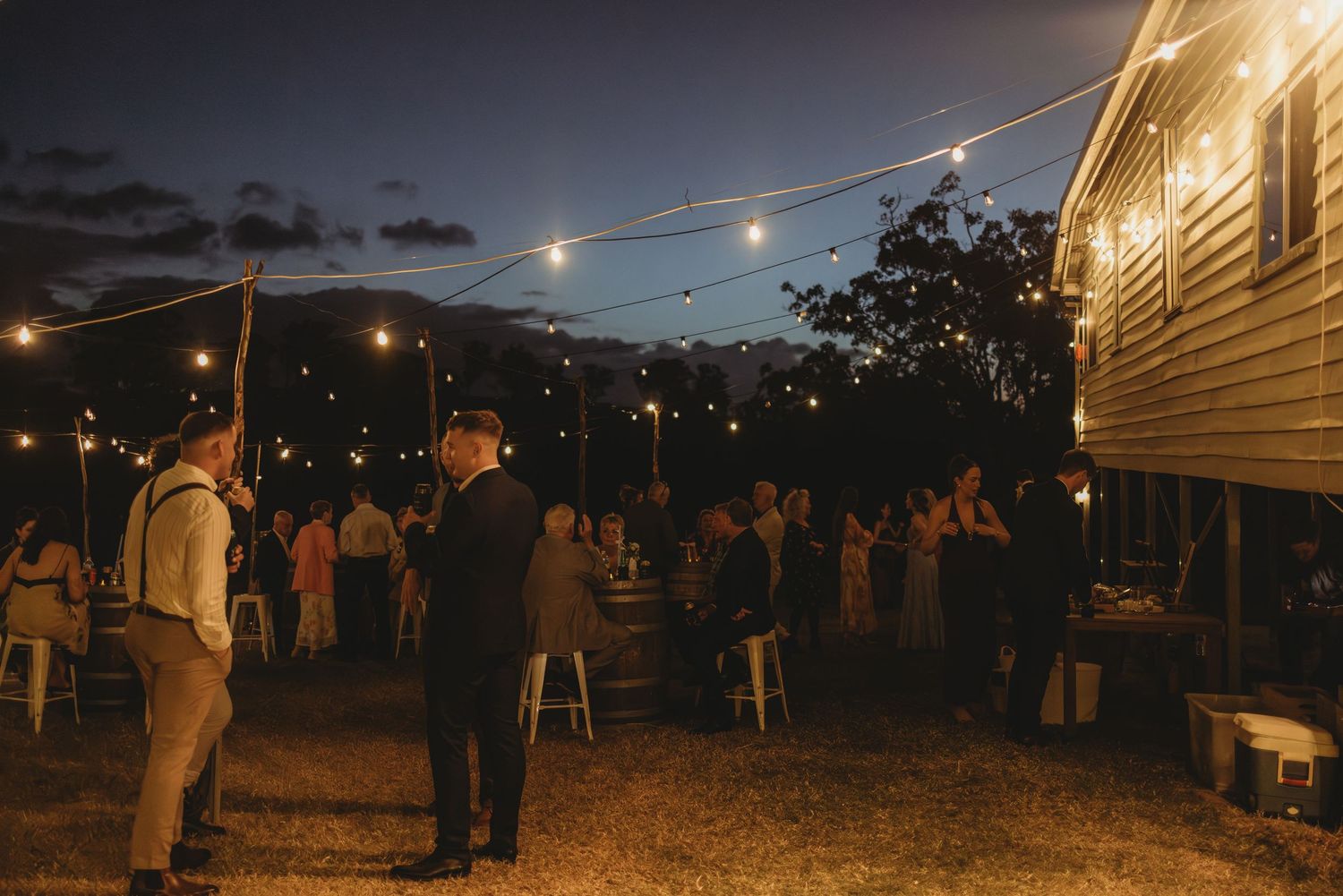 Wedding guests mingle under festoon lights outside rustic venue at twilight reception.