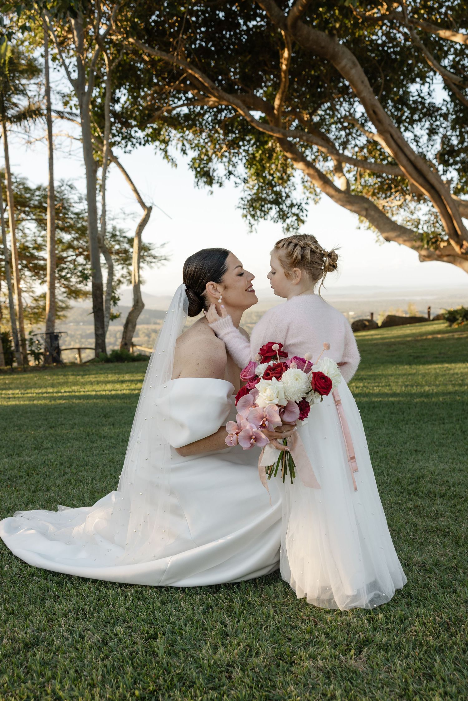 Bride in white gown kneels beside flower girl in pink dress, sharing a tender moment outdoors under a tree.