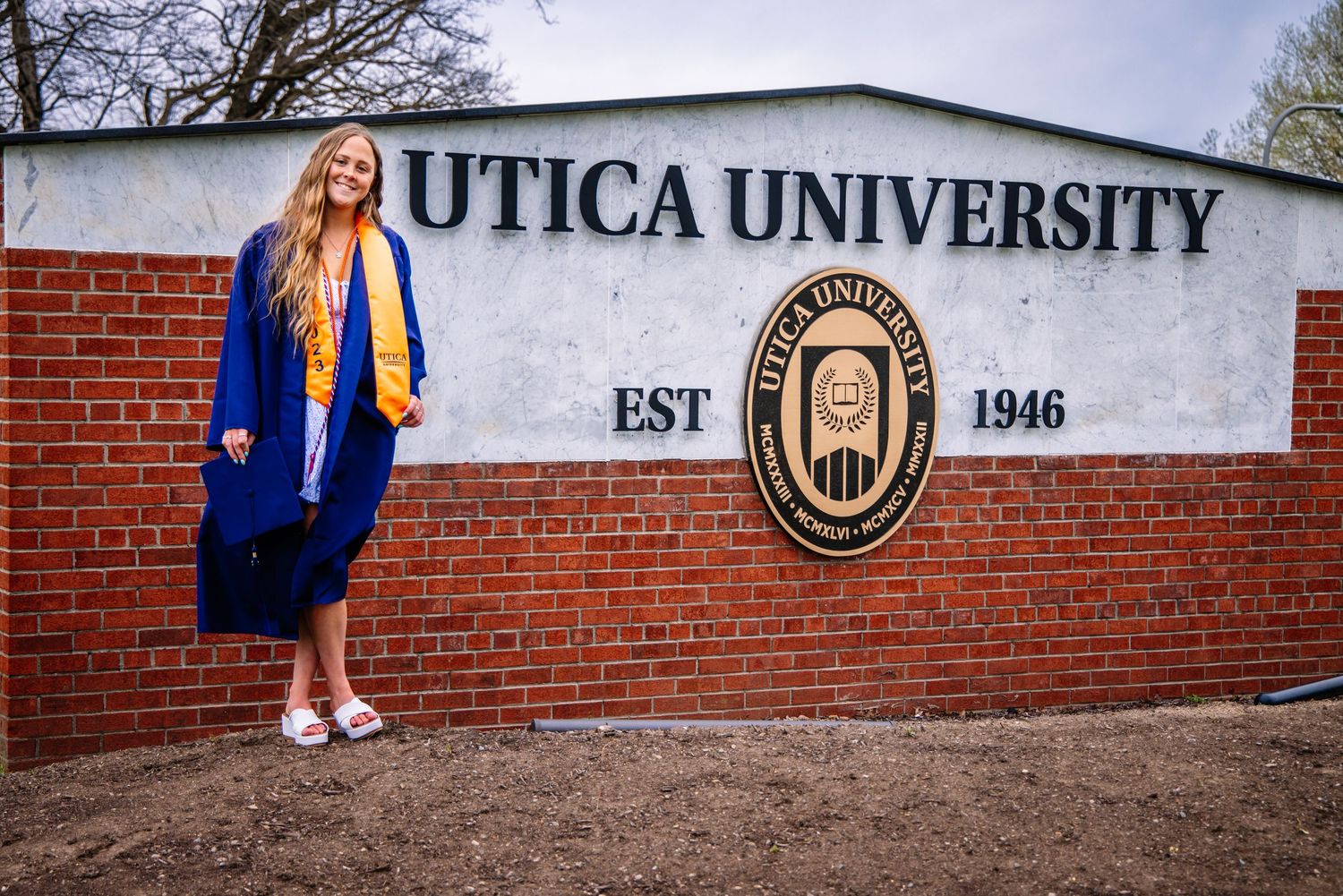 Girl wearing graduation gown in front of Utica University sign