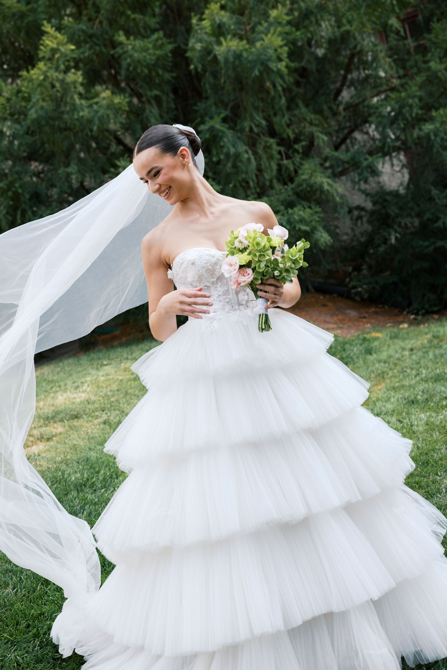 Smiling bride in tiered white tulle ballgown holds floral bouquet outdoors, veil flowing in breeze with green trees behind.