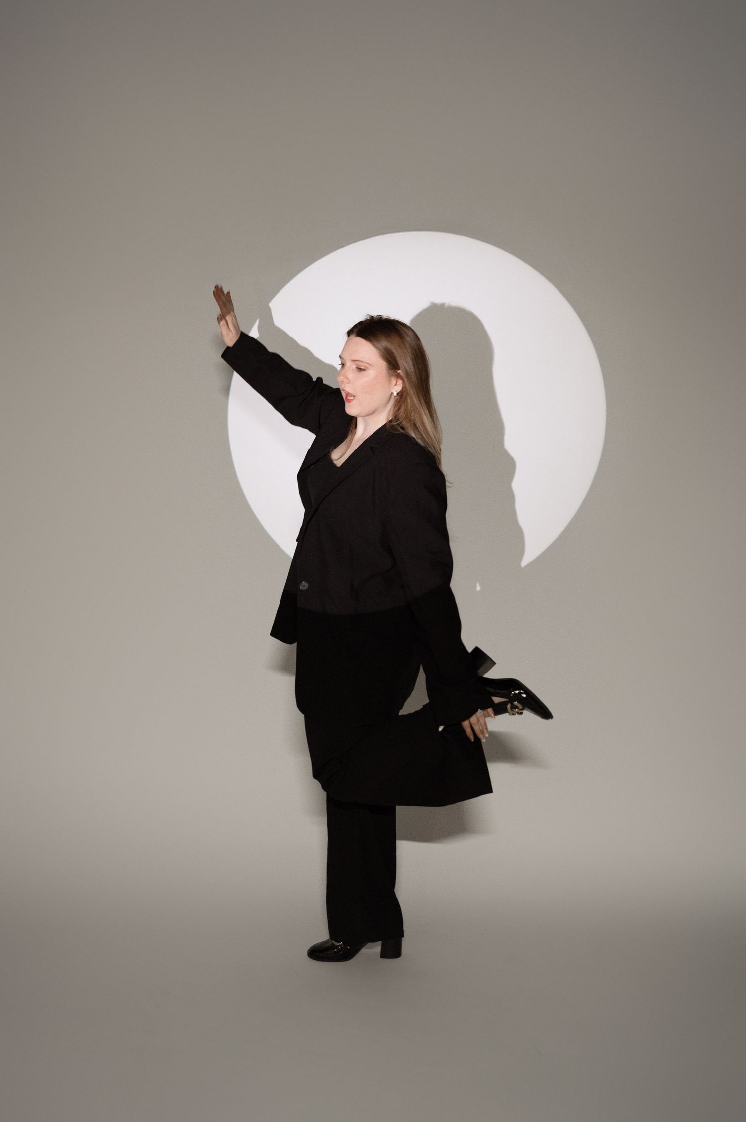 Figure in black suit performs dynamic dance movements against grey background with white circular lighting element.
