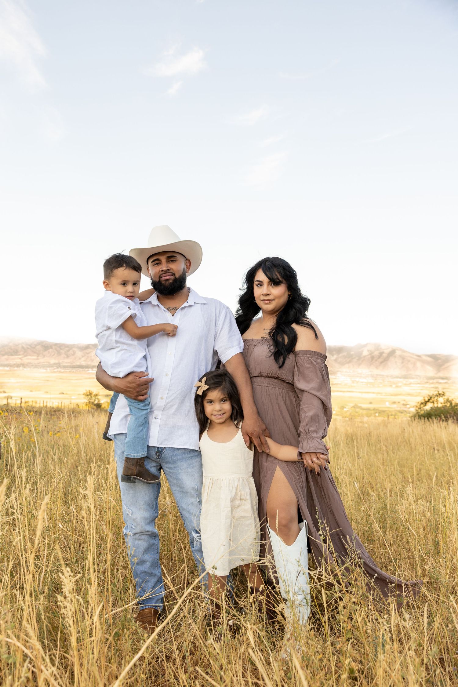 Family of four poses in golden field, father in cowboy hat holds toddler, pregnant mother in mauve dress with daughter.