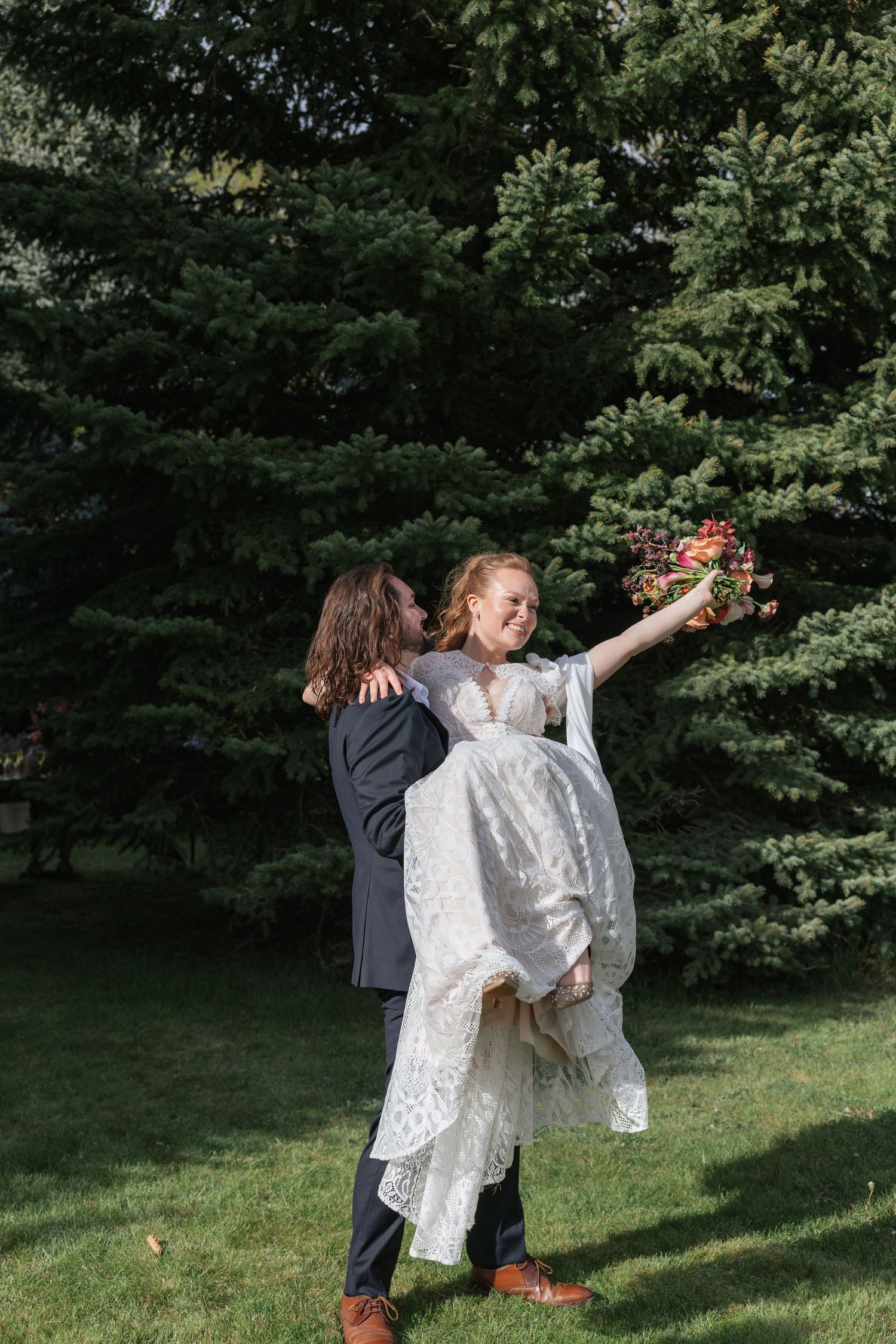 Groom lifting bride in white lace dress as she holds bouquet, celebrating outdoors near evergreen trees.